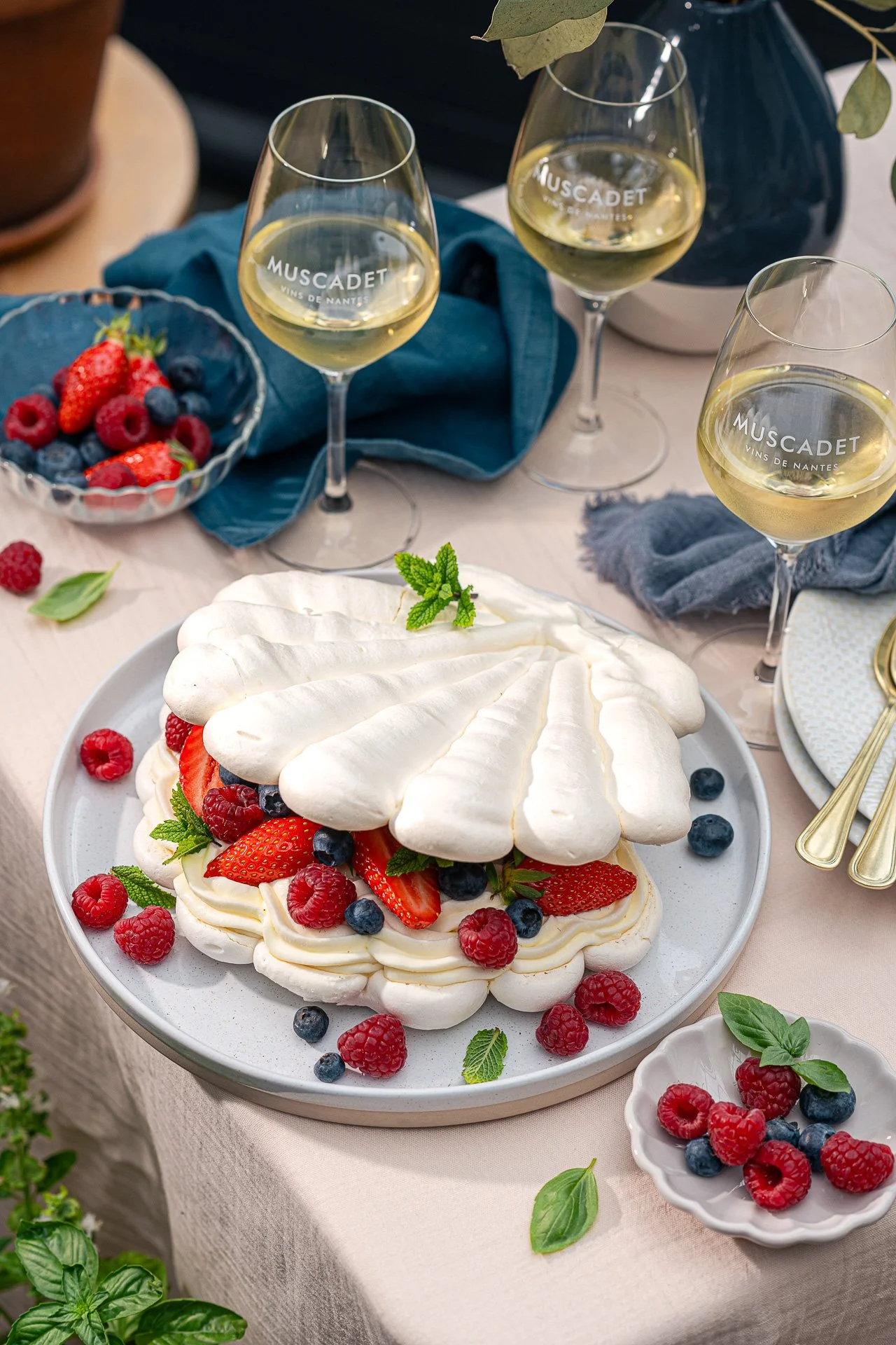 Pavlova meringue aux fruits rouges, avec verres de vin blanc, petits bols de fruits rouges, sur une table avec nappage beige et décorations végétales.