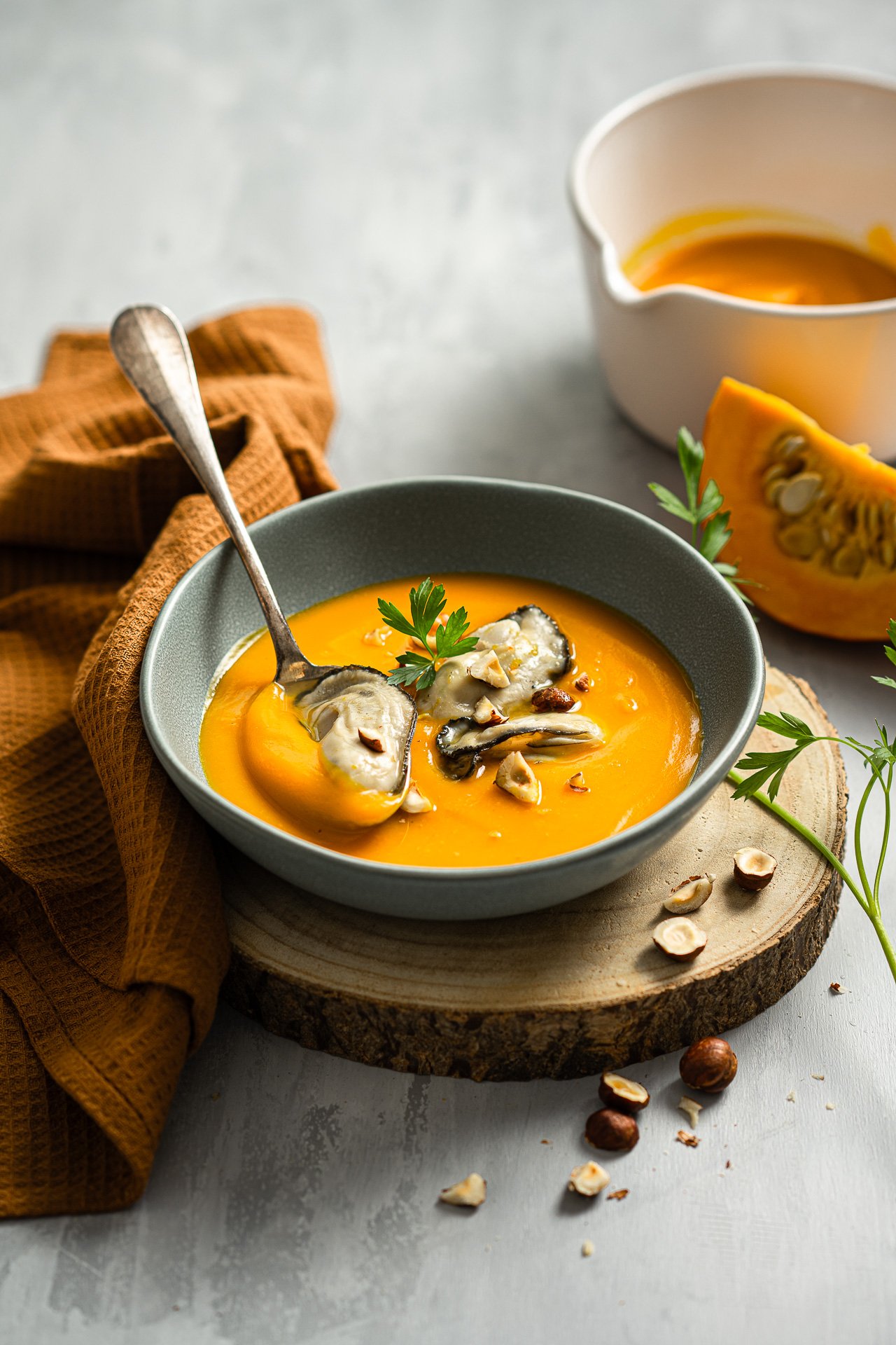 Soupe à la courge butternut garnie de noix et de persil dans un bol gris, avec un morceau de courge, une petite carafe de crème et quelques noix autour.