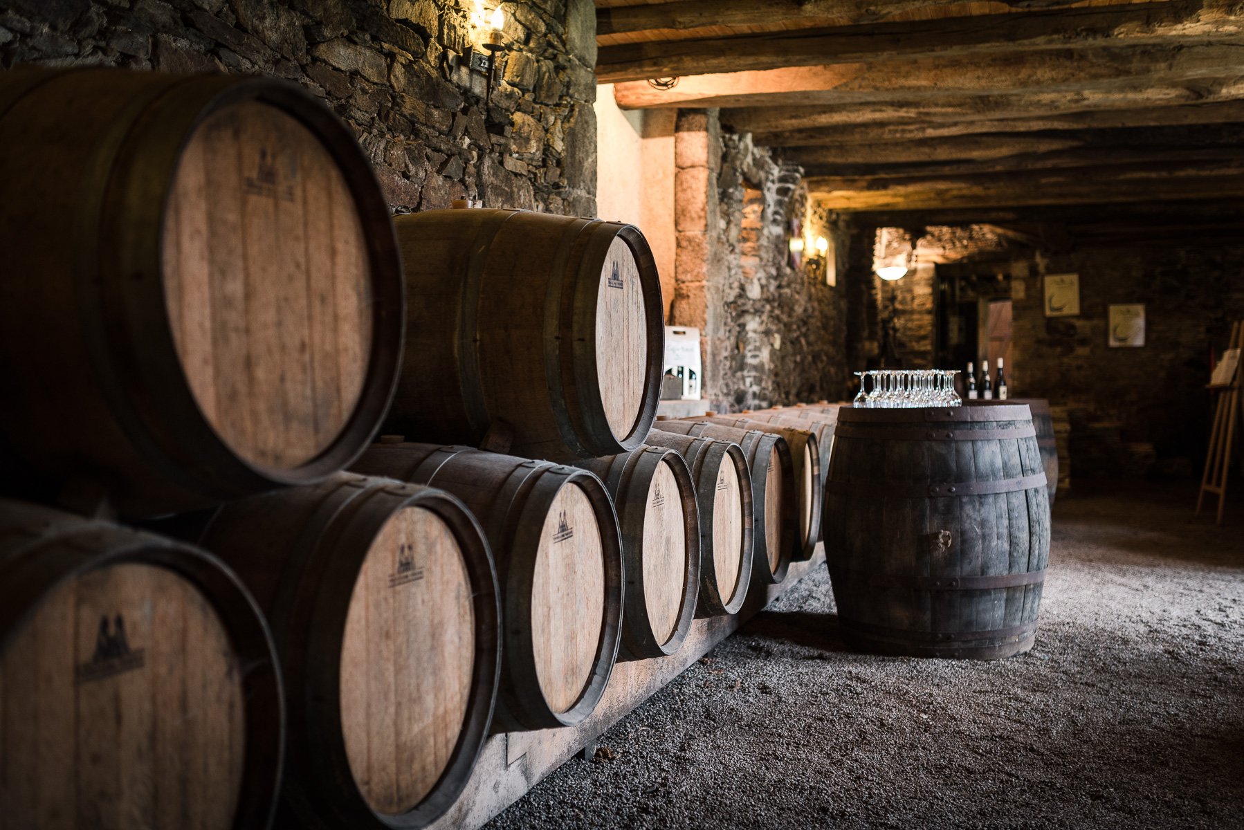 Barils de vin en bois dans une cave en pierre, avec une table faite à partir d'un baril portant des verres à vin et des bouteilles en arrière-plan.