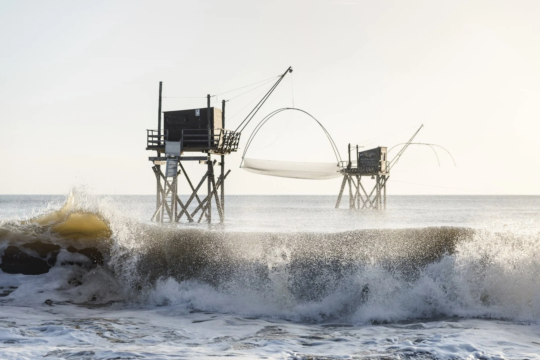 Huit salons de pêche sur pilotis dans la mer, avec des filets de pêche suspendus, sous un ciel clair.
