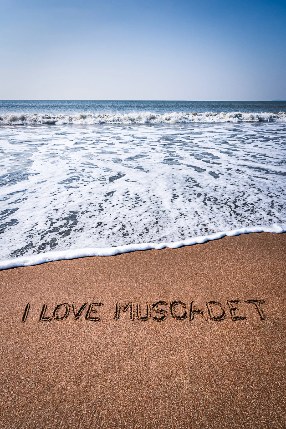 Sable avec écrit en lettres dans le sable : "I LOVE MUSCADE", devant une plage avec des vagues et la mer.