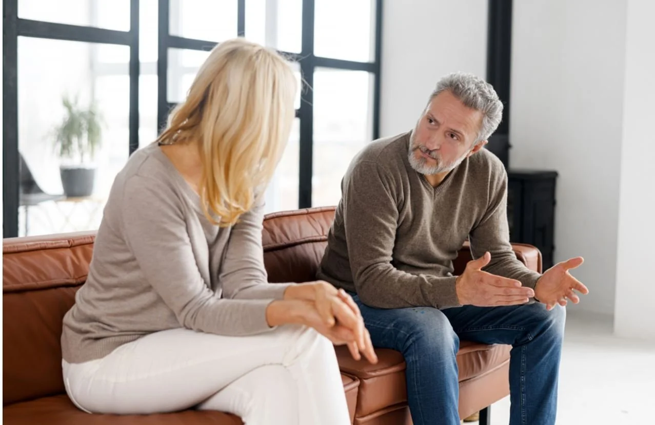 A man and woman sitting on a brown leather couch having a serious conversation, with the man gesturing with his hands.