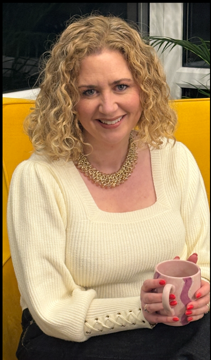 A woman with curly blonde hair, wearing a cream-colored sweater and a pearl necklace, holding a pink mug with purple accents, sitting on a yellow seat with a plant and window in the background.