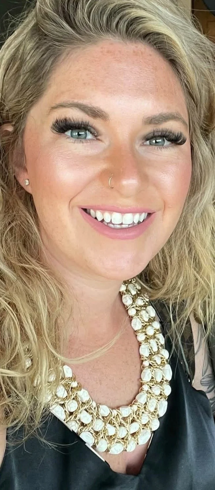 Close-up of a woman with wavy light brown hair, blue eyes, and hoop earrings, smiling slightly at the camera.