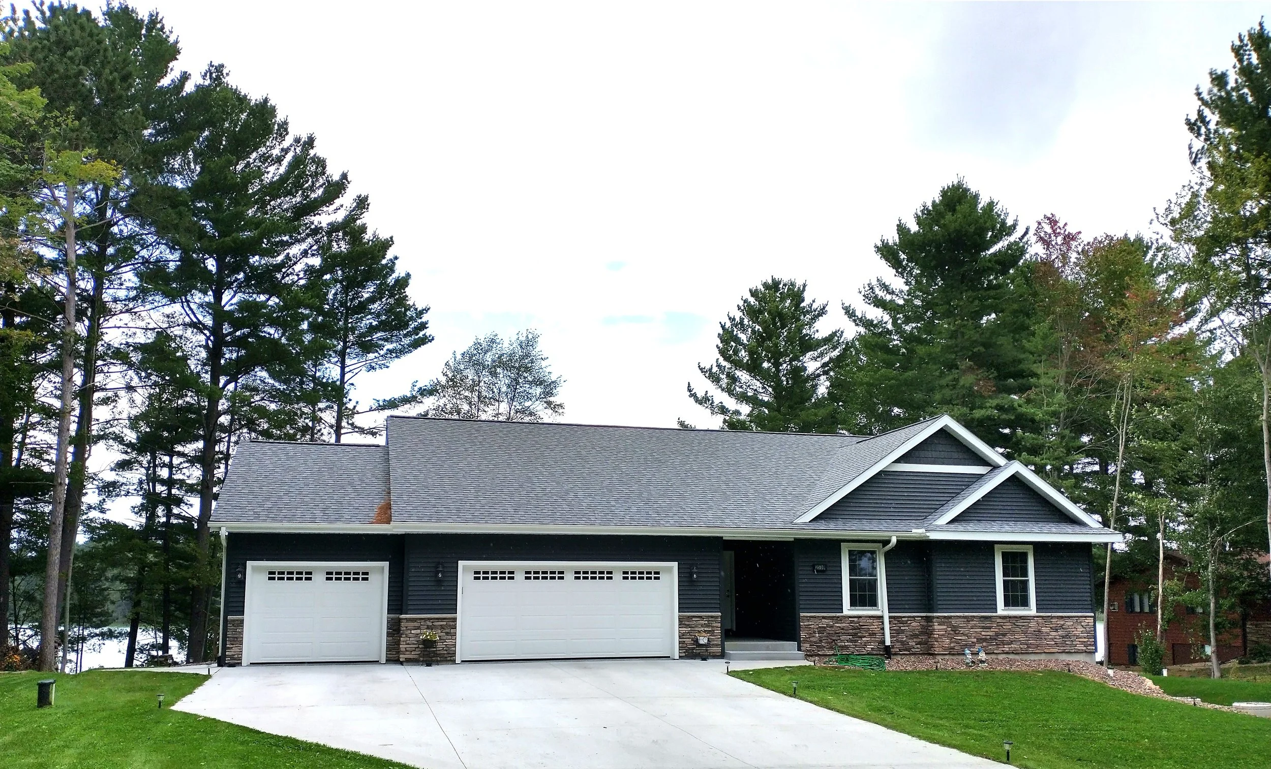 Modern dark gray house with white garage doors and stone accents, surrounded by trees and green grass.