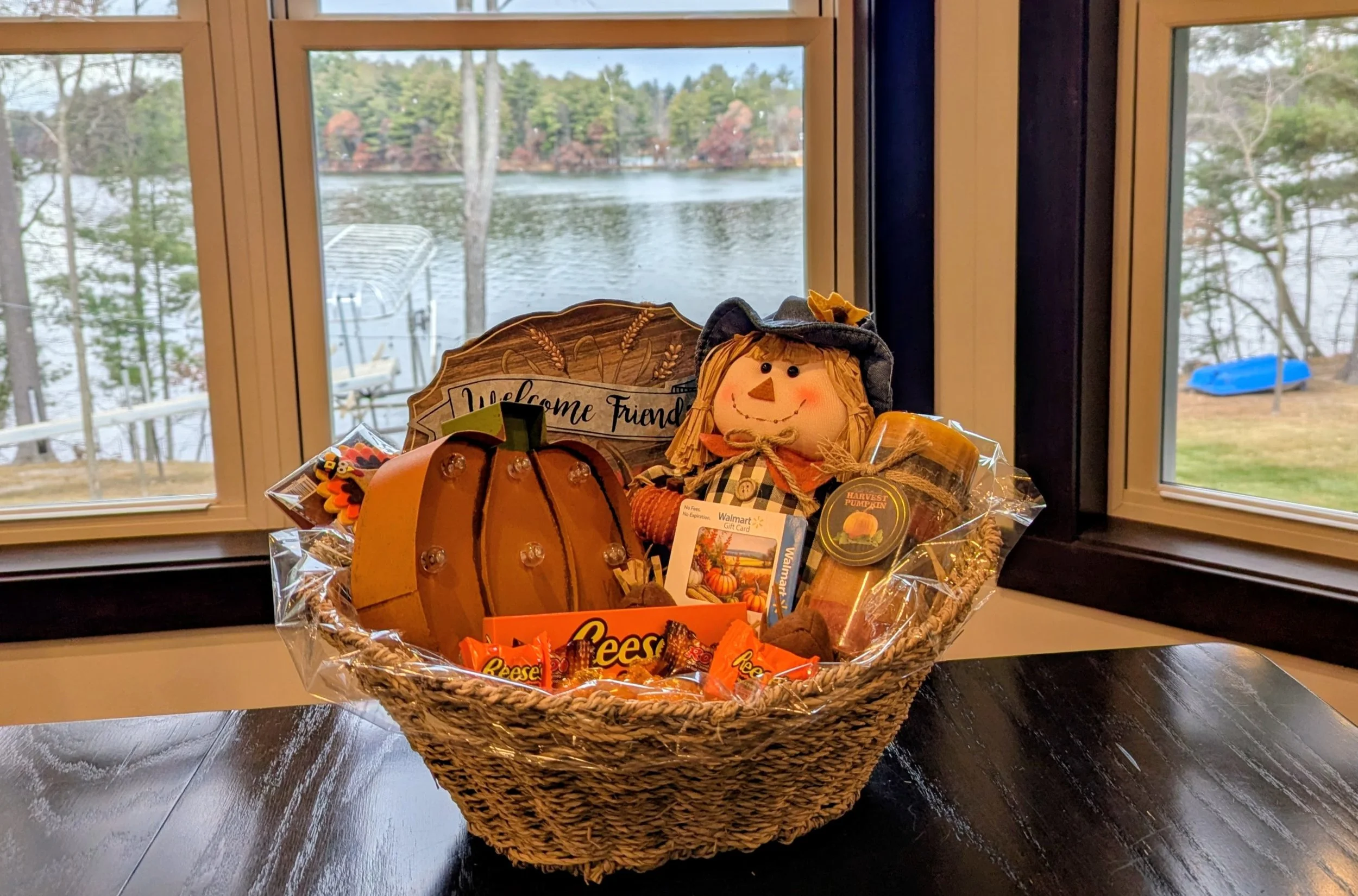 A fall-themed gift basket with a scarecrow doll, pumpkin decor, Reese's candies, and a sign that says "Welcome Friends" on a dark wooden table by a window overlooking a lake with trees.