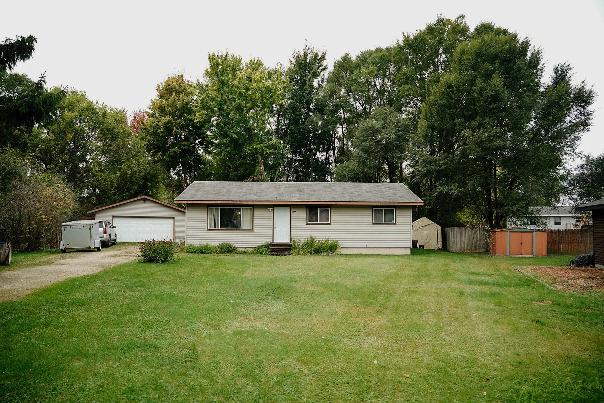 Front view of a beige house with a gray roof, three windows, and a white door, surrounded by green lawn and trees, with a driveway on the left.