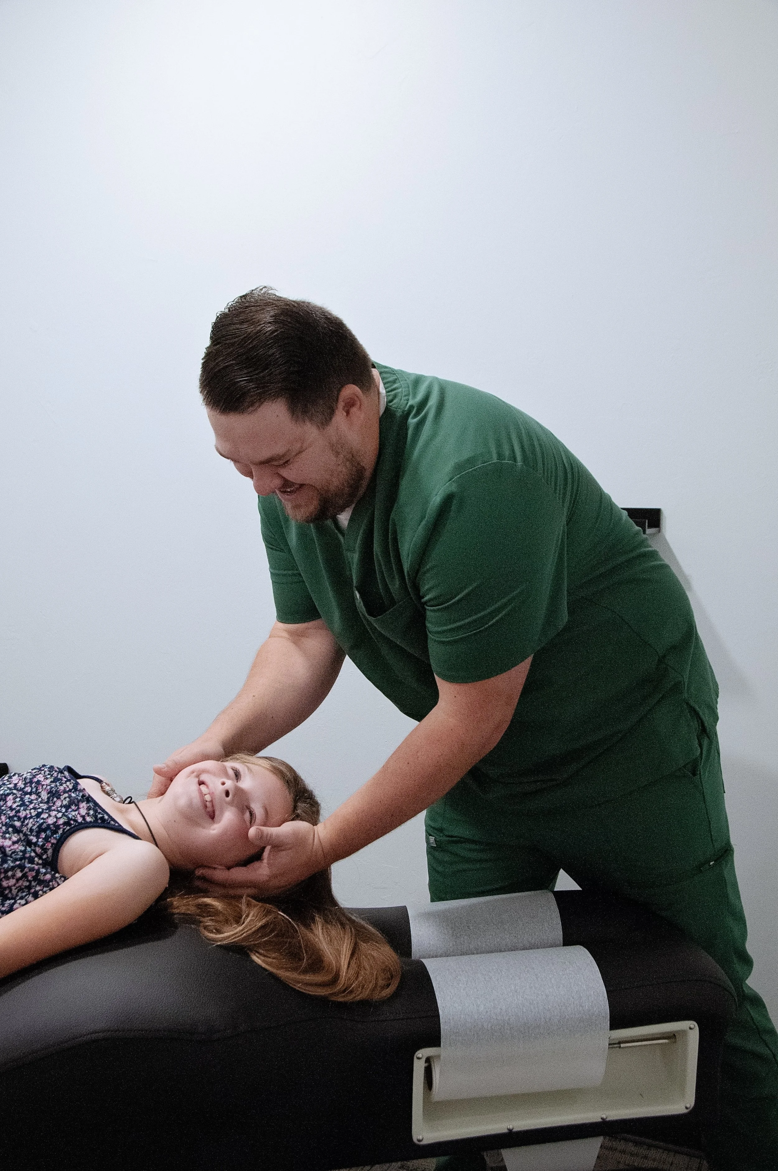 A healthcare professional in green scrubs treating a young girl lying on an examination table, smiling and appearing happy.