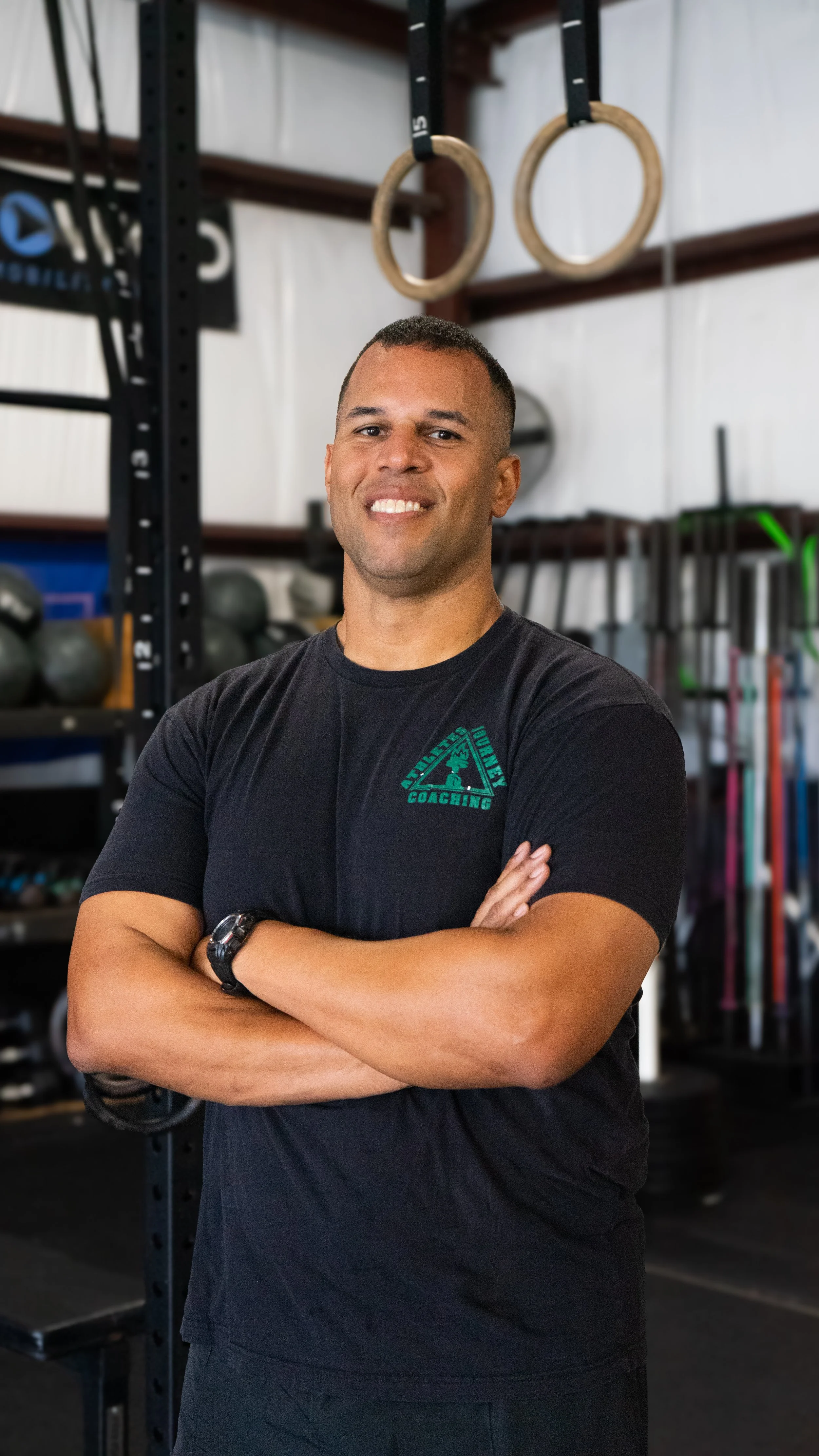 A man standing with arms crossed inside a gym, wearing a black t-shirt with a logo that says "Movement Coaching" and gym equipment like rings and medicine balls in the background.