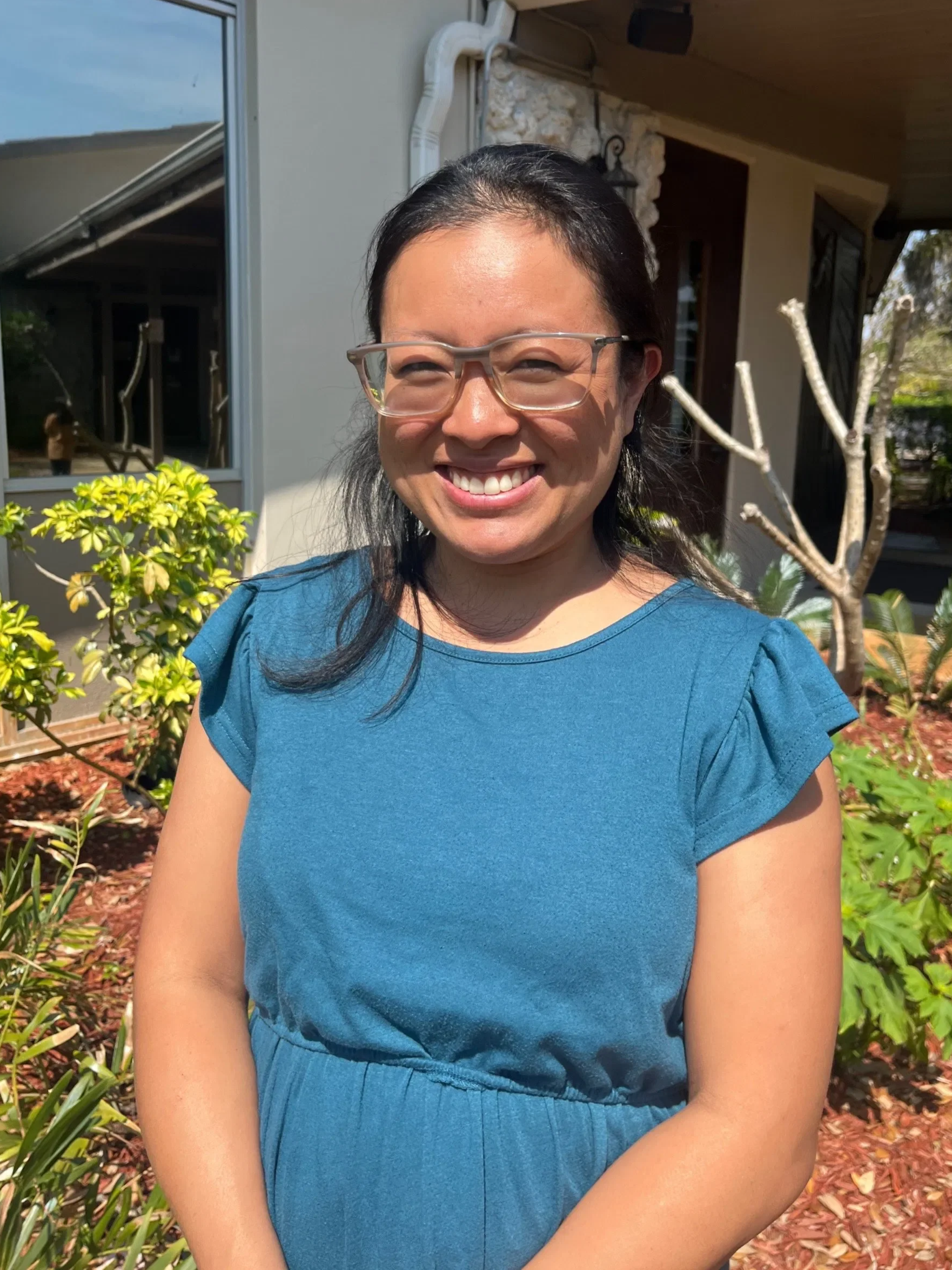 Melinda wearing glasses smiling outdoors, wearing a blue top, with plants and a house in the background.