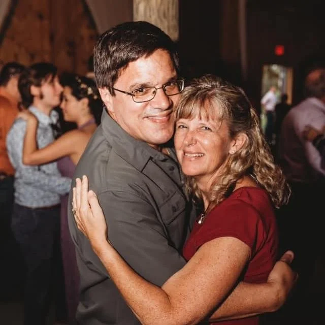 A smiling middle-aged couple dancing closely at a social event, with other couples dancing in the background.