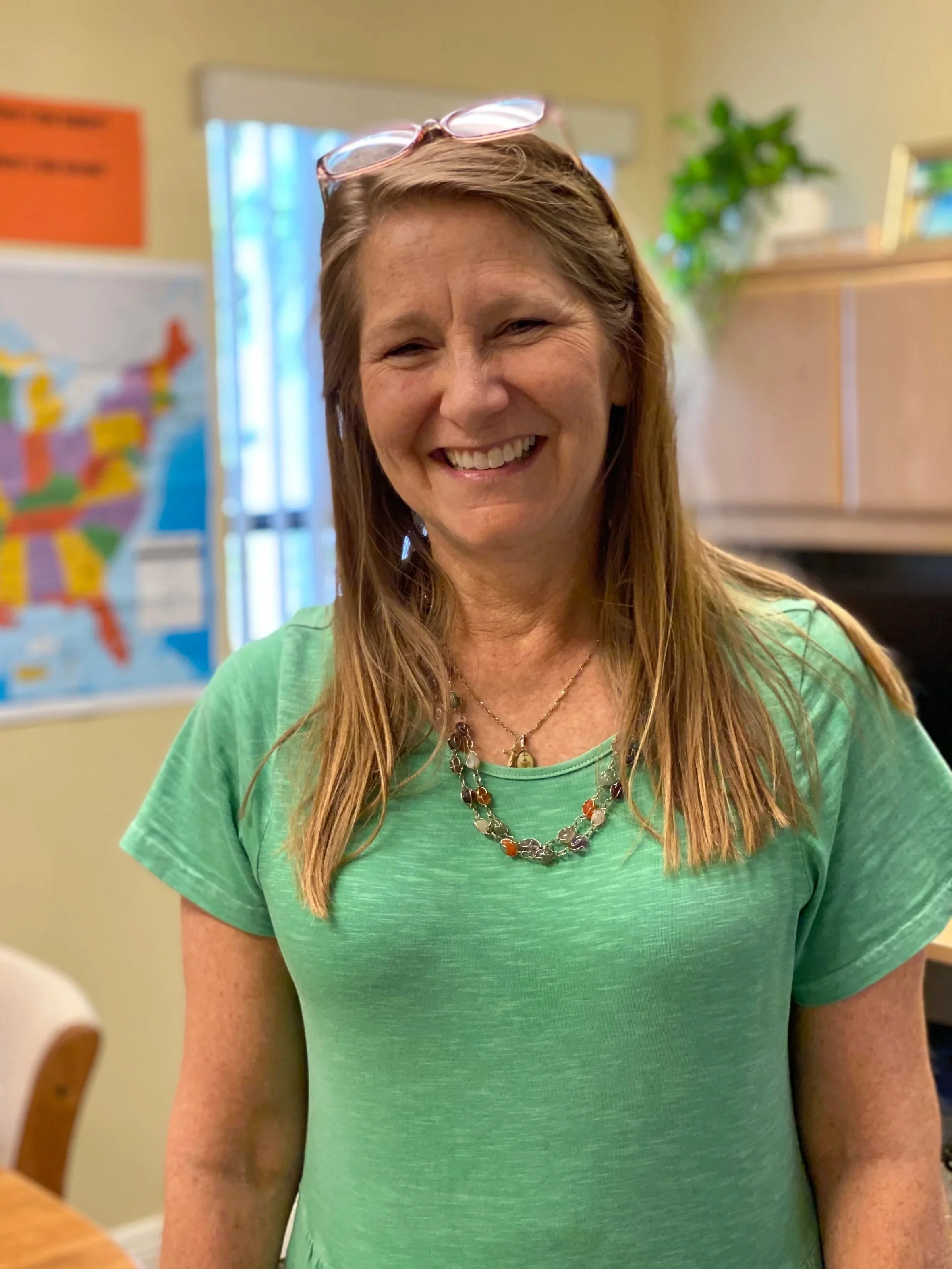 Julie in a green t-shirt, and multiple necklaces, smiling at the camera inside a room with a map of the United States on the wall and a plant in the background.