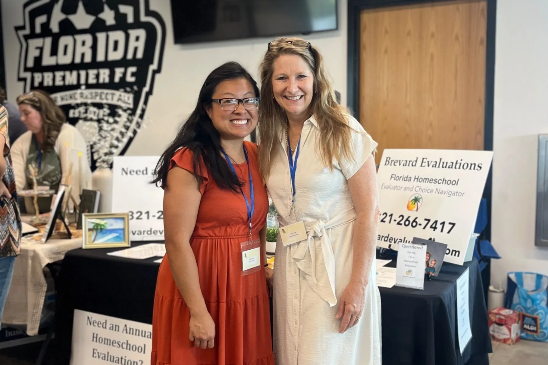 Two women standing and smiling at a conference or event, with booths and signs behind them. One woman is wearing an orange dress and glasses, while the other is in a cream-colored dress.