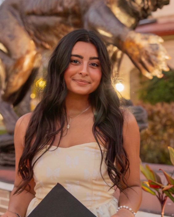 Alina smiling outdoors with a large metal sculpture behind her and warm lighting.