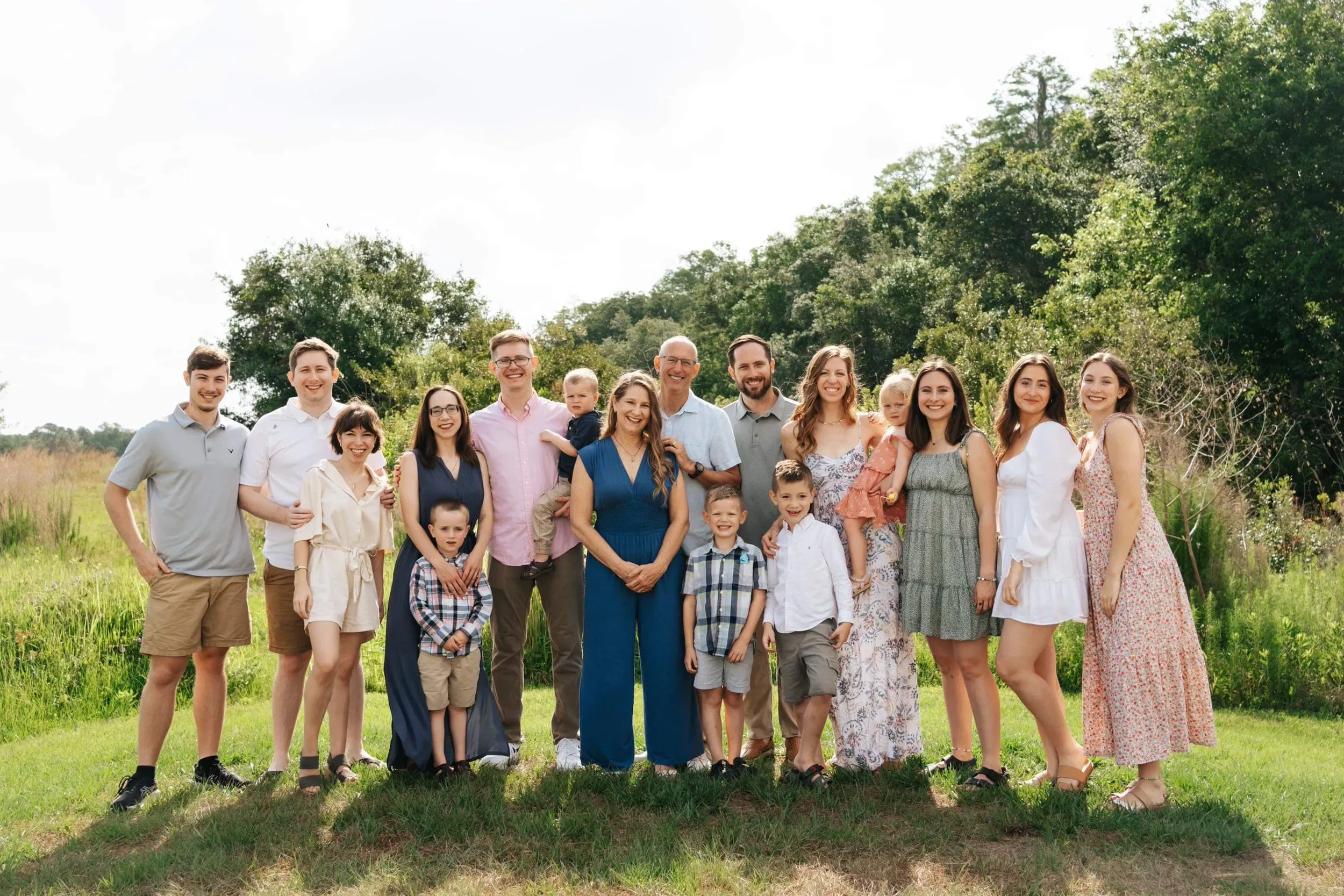 A large multi-generational family group standing outdoors in a grassy field with trees in the background, smiling and posing for the camera.