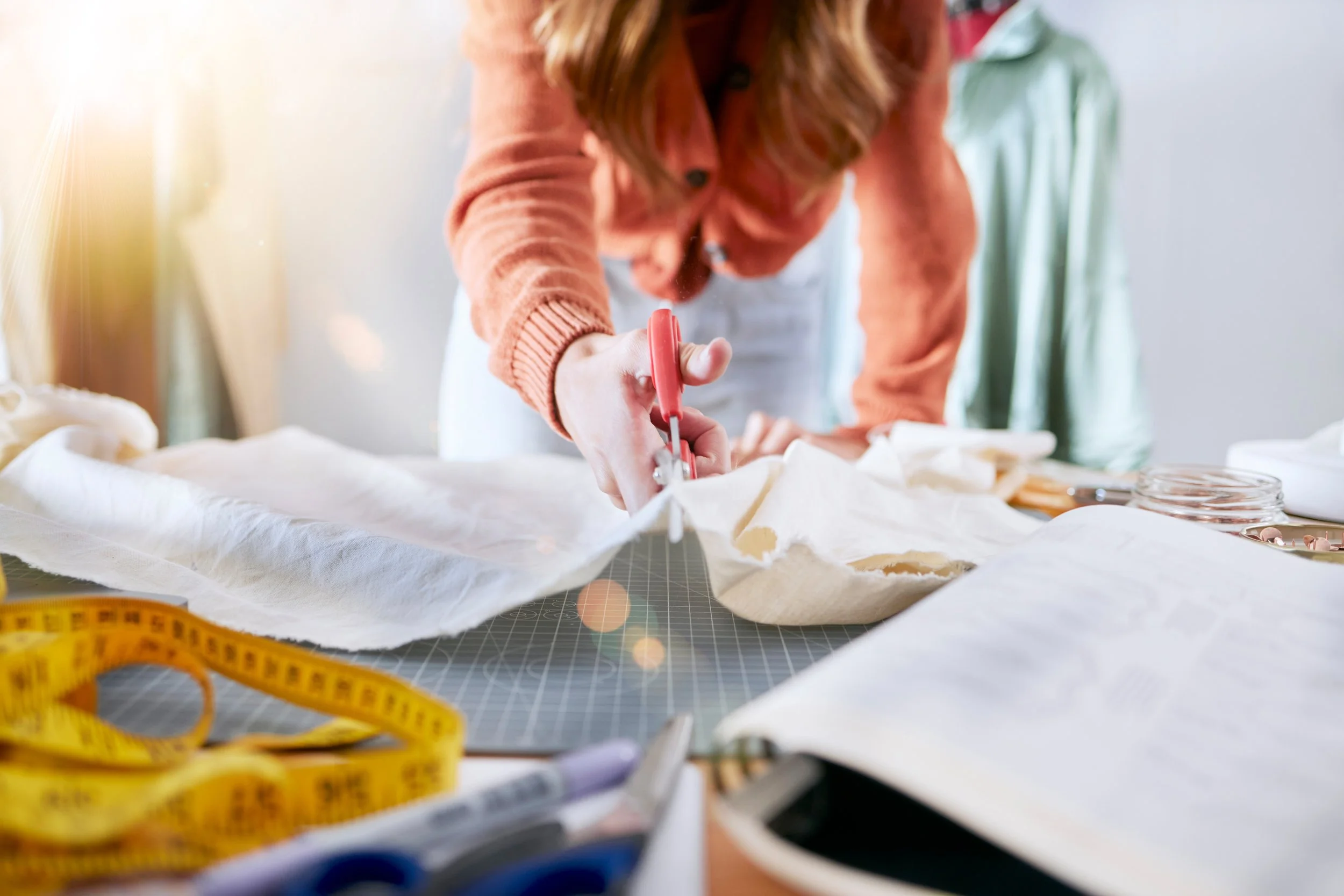 A woman using scissors to cut fabric or paper on a table with sewing or design supplies, including a yellow measuring tape and jars, in a well-lit room.