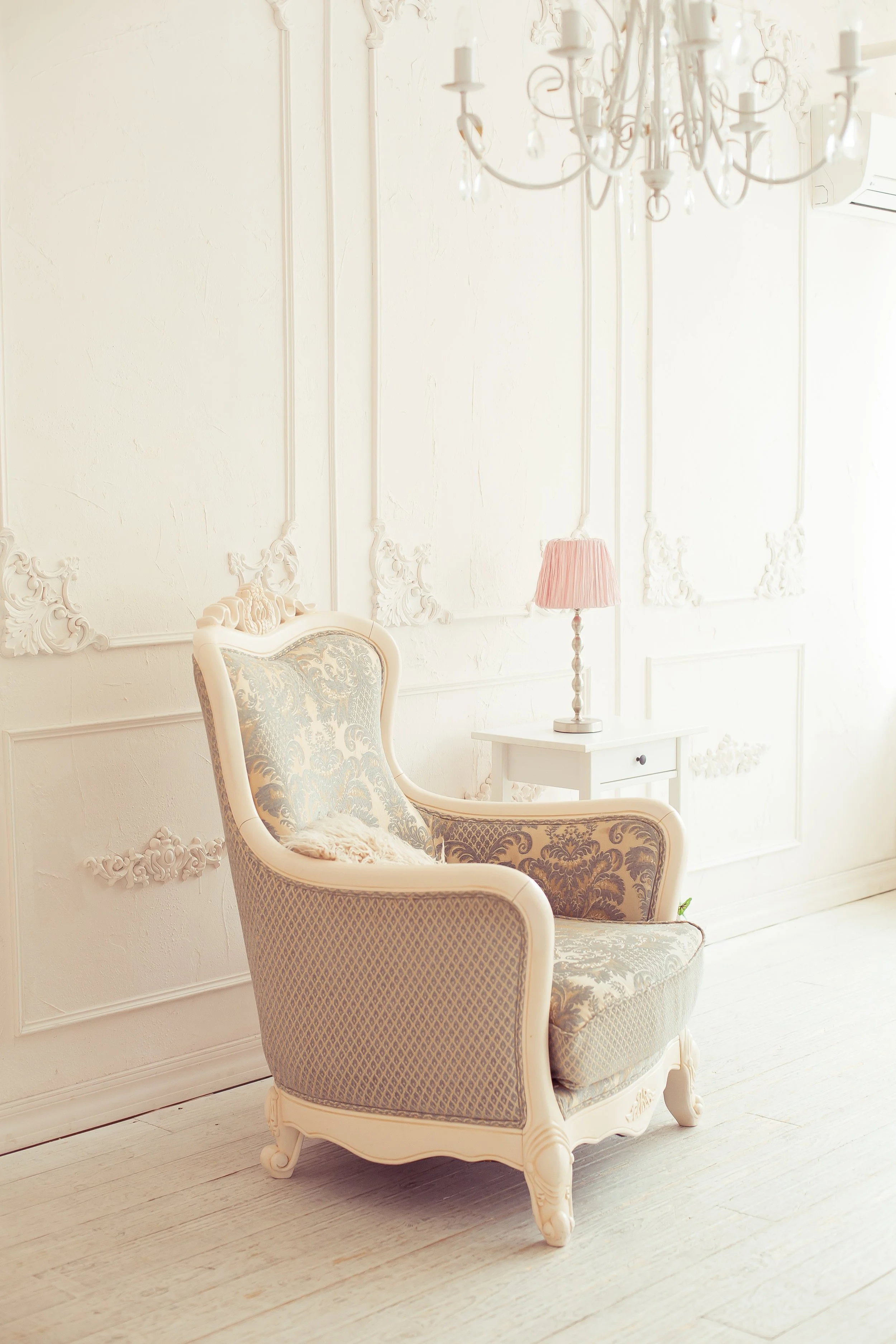 Elegant vintage armchair with floral upholstery and ornate carved wooden frame in front of a white wall with decorative molding, beside a small white side table with a pink table lamp and an air conditioning vent, on a light-colored wooden floor.