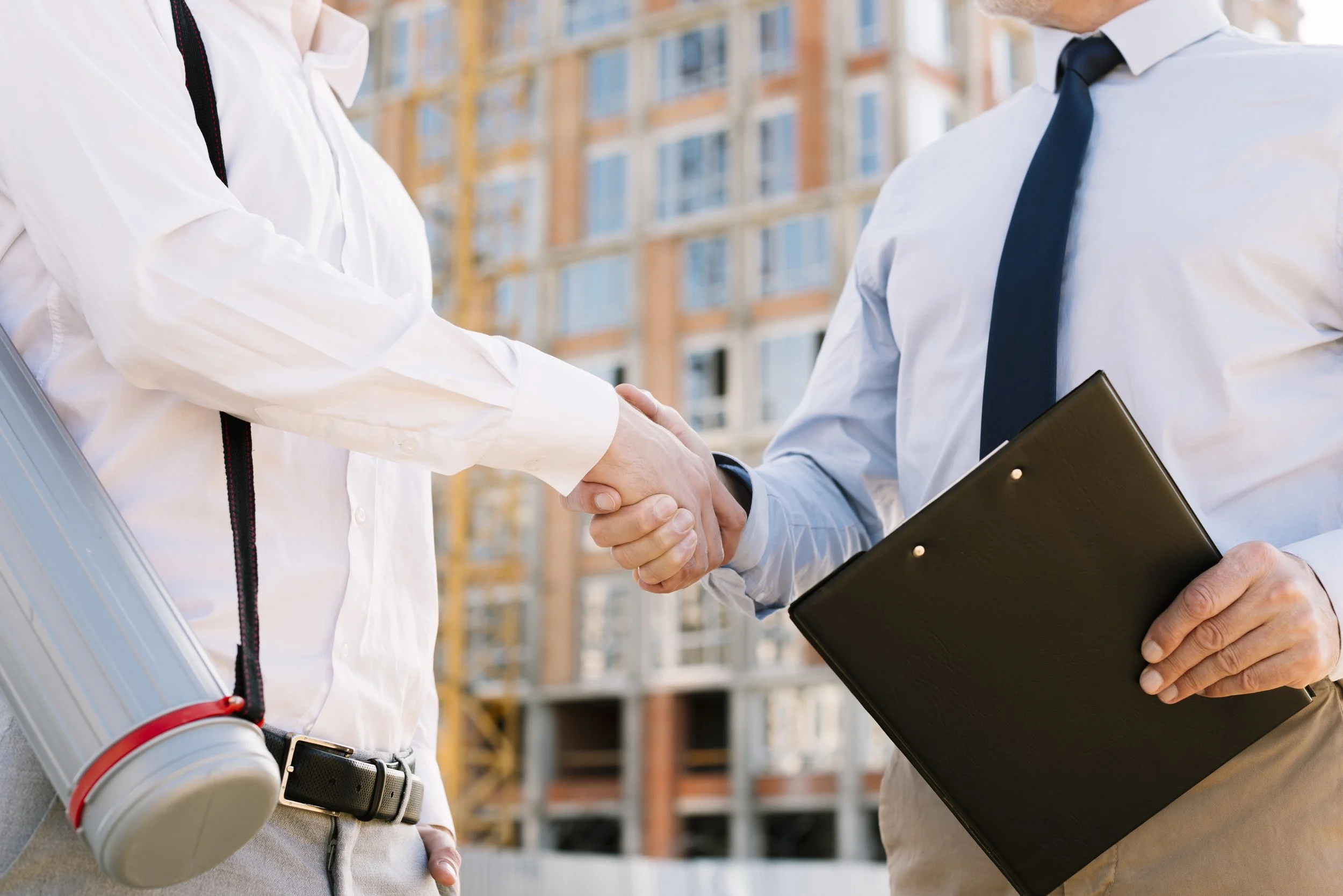 Two people shaking hands in front of a construction site, one of them holding a clipboard.