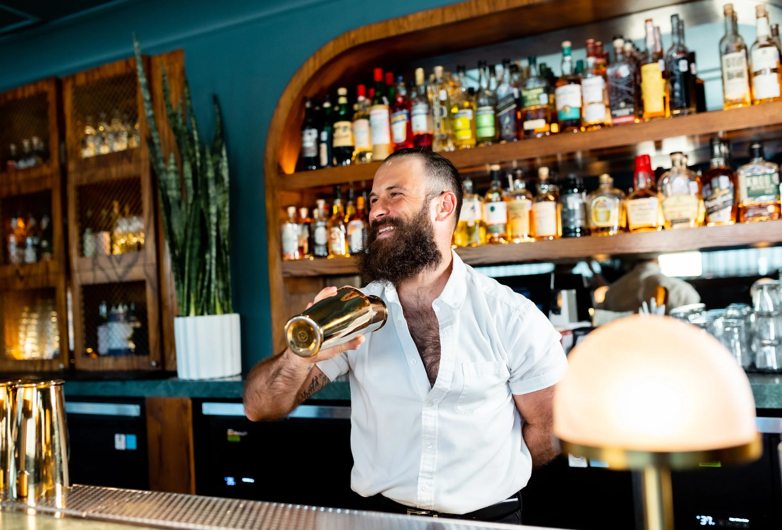 A bearded man in a white shirt smiling and holding a gold cocktail shaker behind a bar with liquor bottles on shelves in the background.