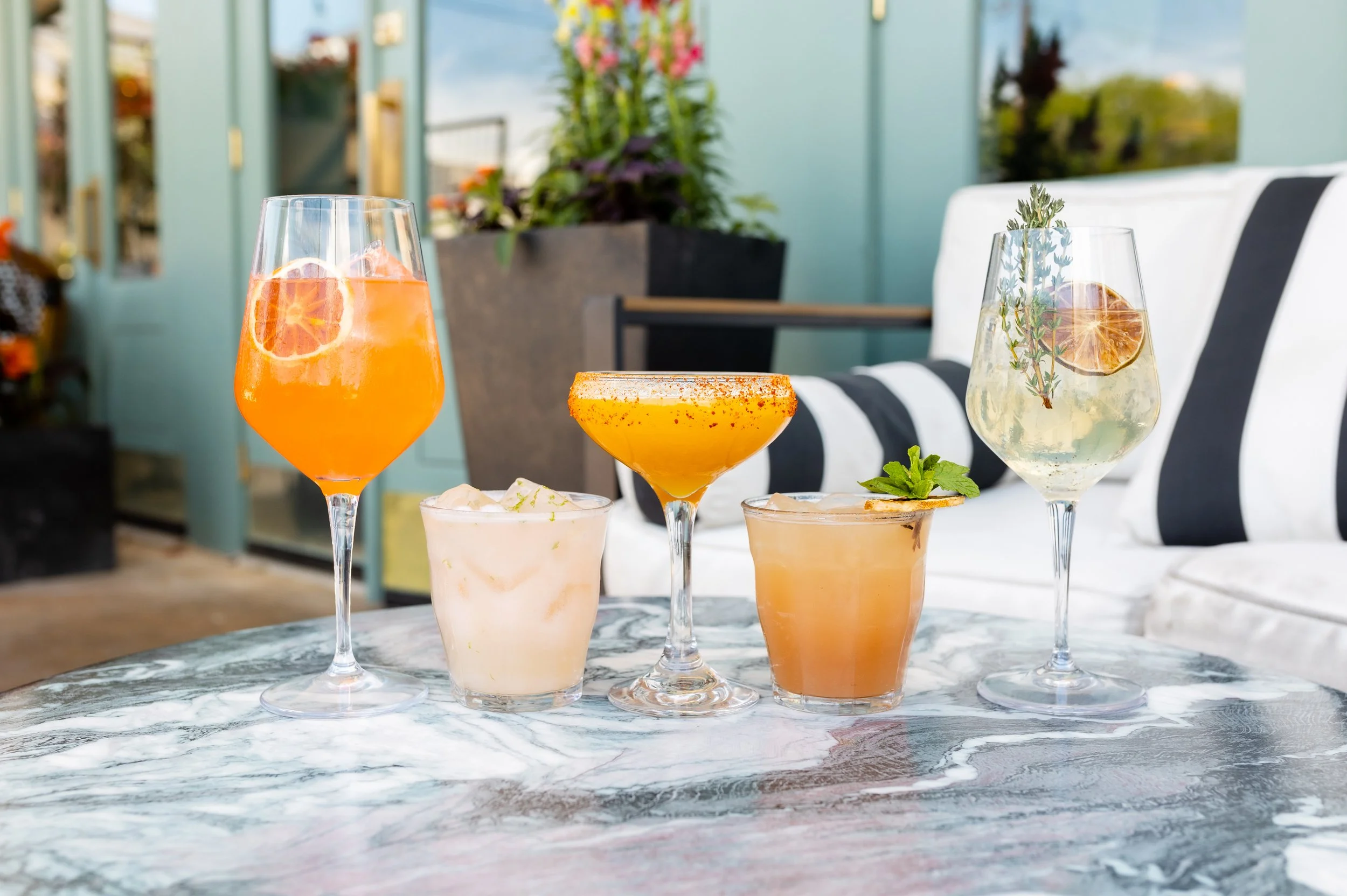 A collection of five colorful cocktails with garnishes on a marble table, with a patio background including cushions and plants.