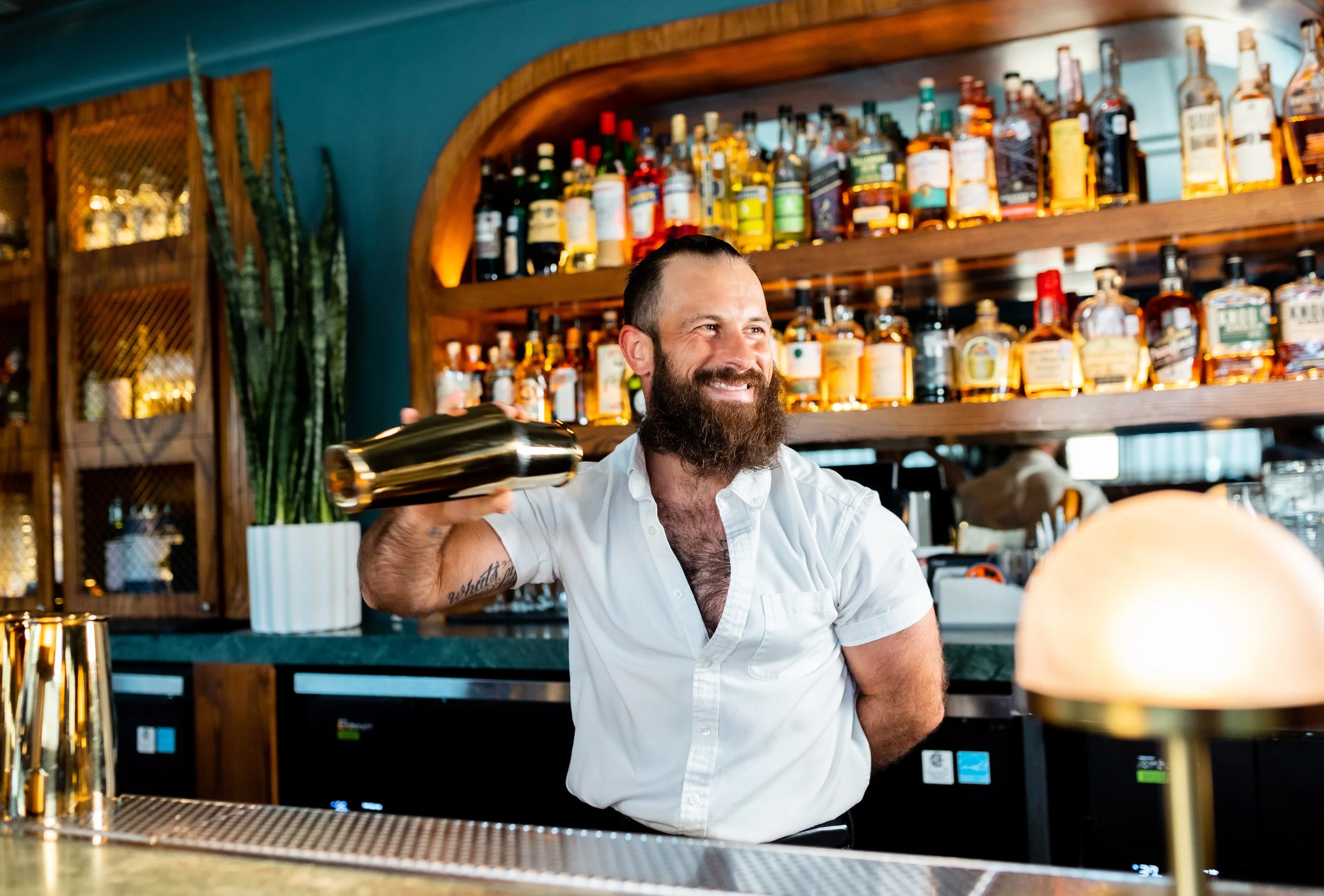 A smiling bartender with a beard, wearing a white shirt, shaking a cocktail shaker at a bar with liquor bottles on shelves behind him.