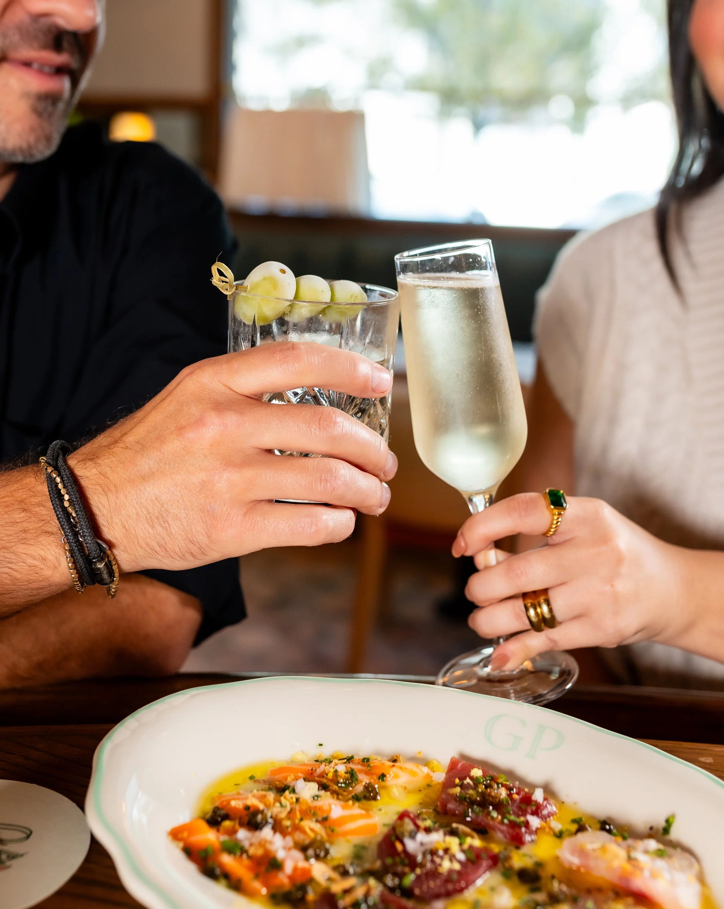 Two people toasting with champagne & a cocktail, with a plate of food below, in a restaurant setting.