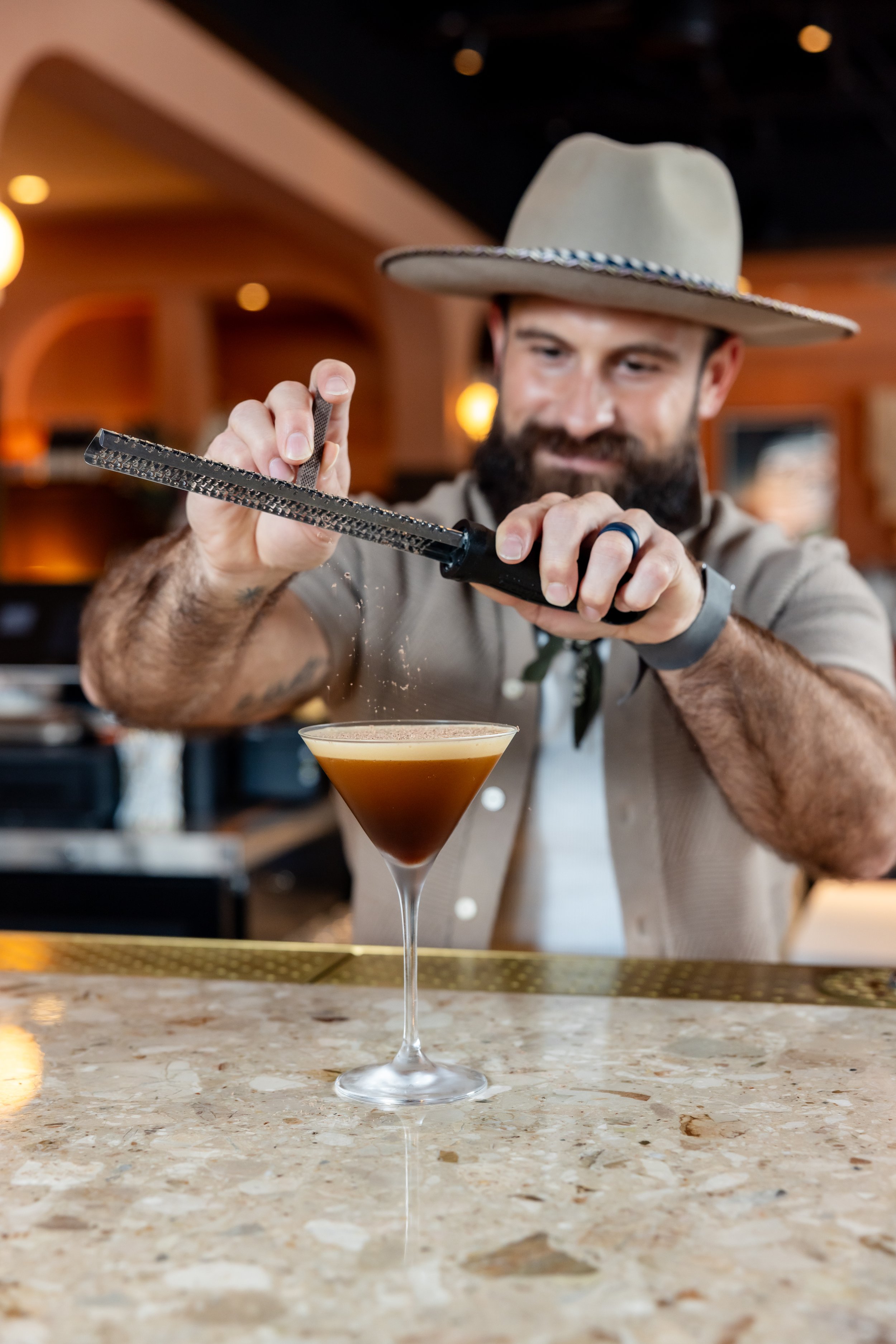 A man with a beard wearing a beige cowboy hat and a beige shirt is using a grater to add shaved chocolate to a cocktail on a bar counter.
