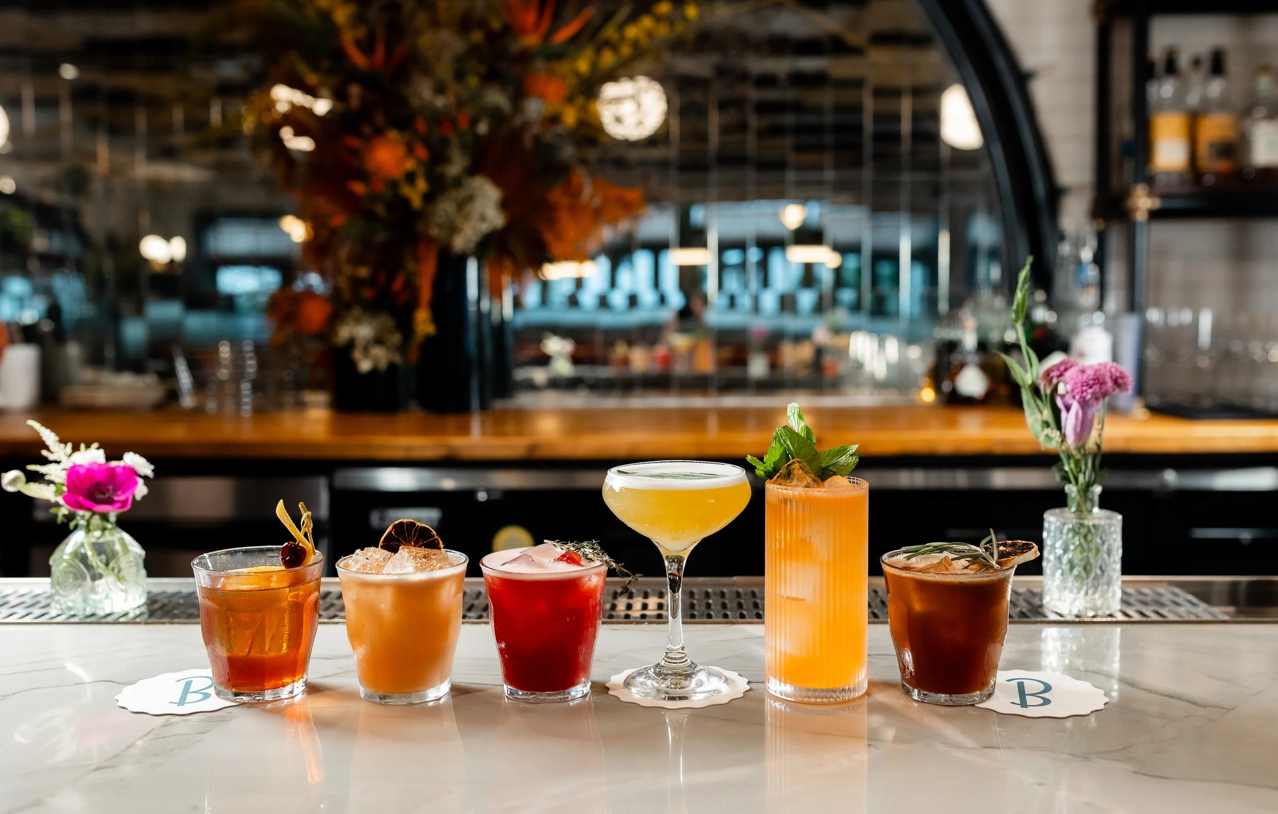 Six colorful cocktails arranged on a bar counter with flowers in vases in the background.