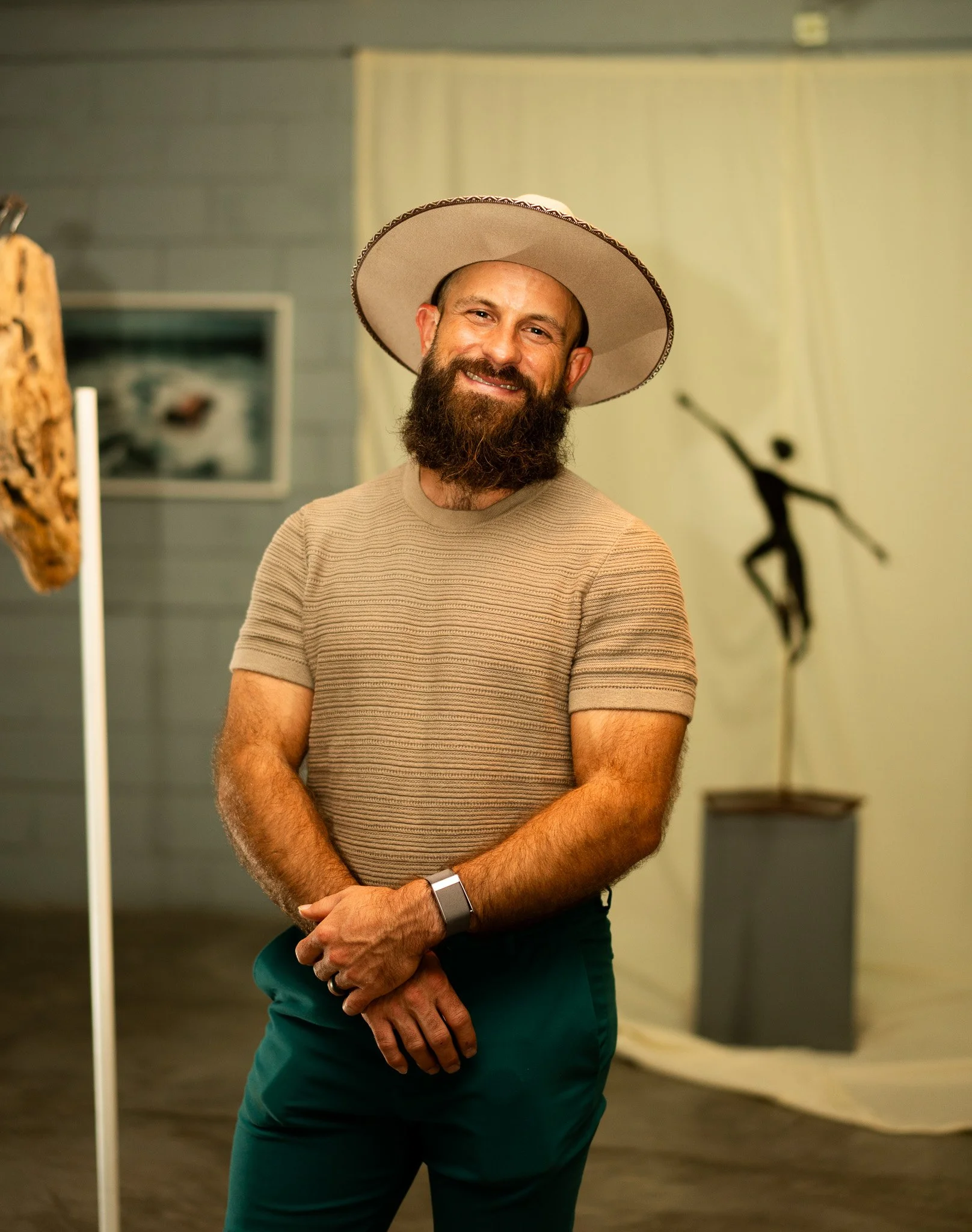 A man with a beard wearing a wide-brimmed hat, a beige striped t-shirt, teal pants, and a smartwatch, smiling and standing indoors in front of a sculpture of a dance figure on a pedestal and a painting.