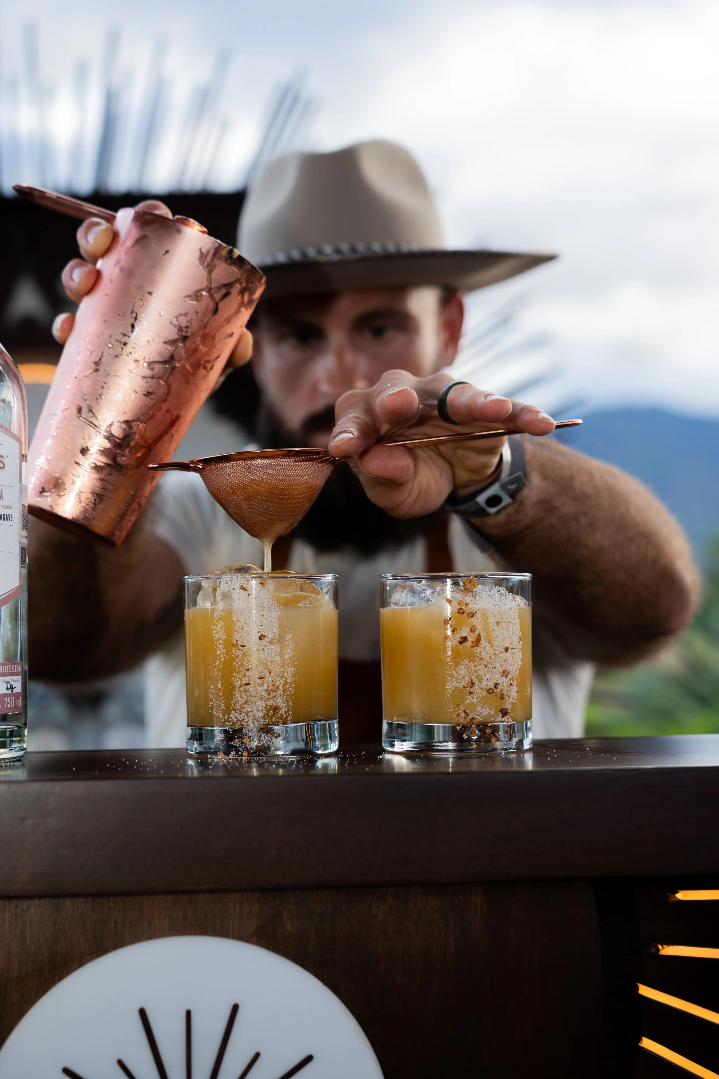 A bartender in a white shirt and wide-brimmed hat preparing cocktails with a copper shaker and strainer, pouring liquid into two glasses filled with orange-colored drinks, on a bar counter with a scenic outdoor background.