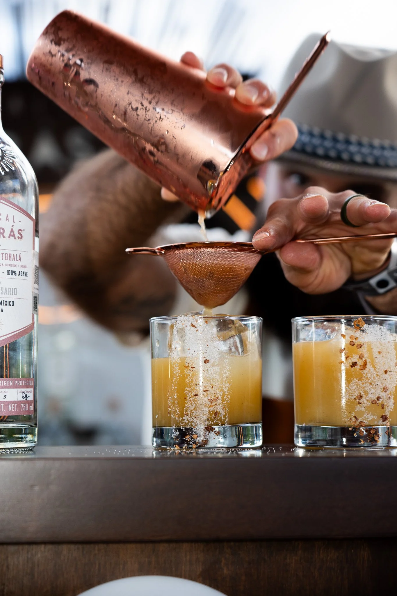 A bartender pouring a liquid through a strainer into a glass with lime or lemon in it, with another similar drink beside.