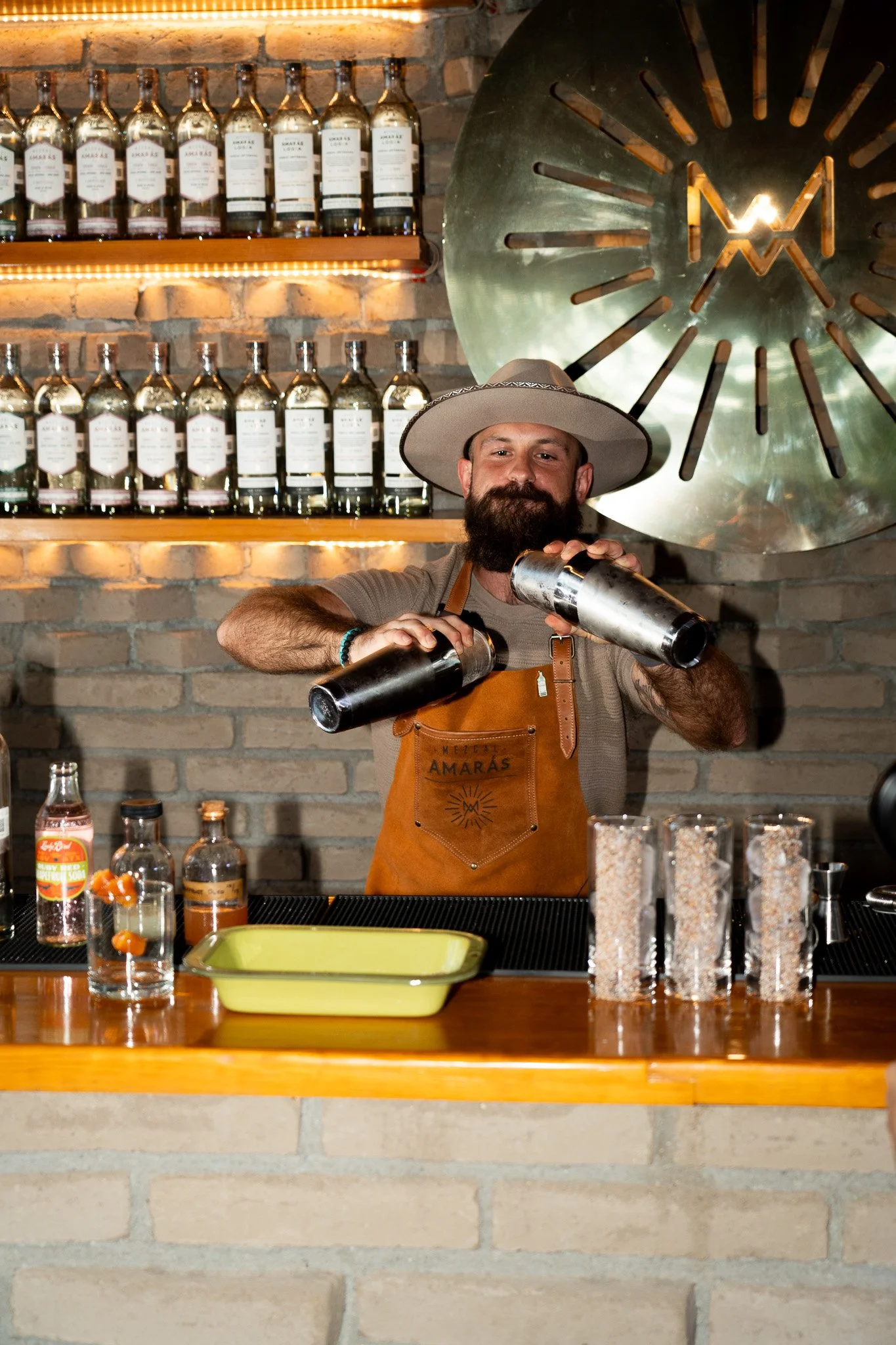 Bartender with a beard and hat shaking cocktails behind a bar with bottles and glasses.