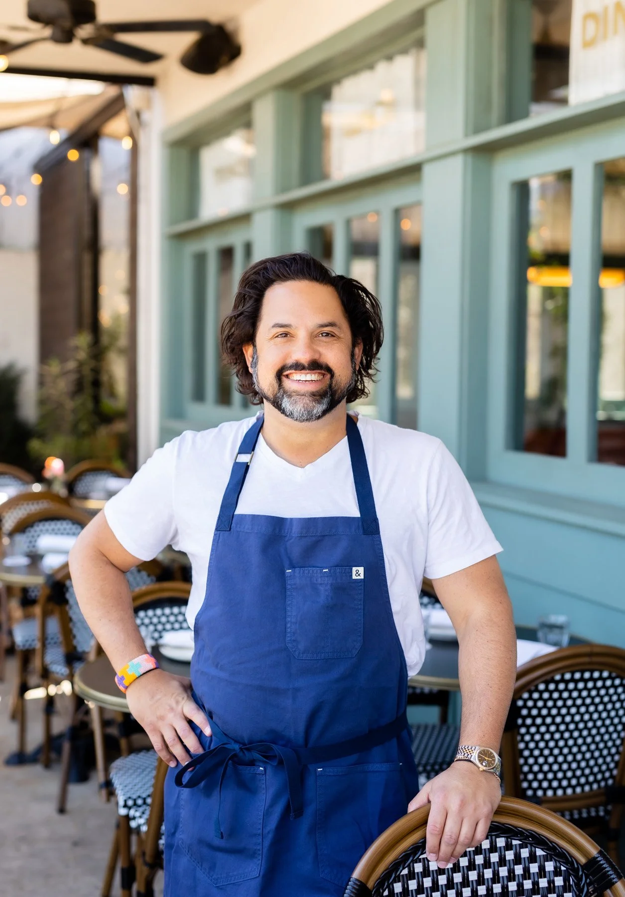 Smiling man with dark wavy hair and beard wearing a white t-shirt and a blue apron, standing outside of a restaurant or cafe with outdoor seating.