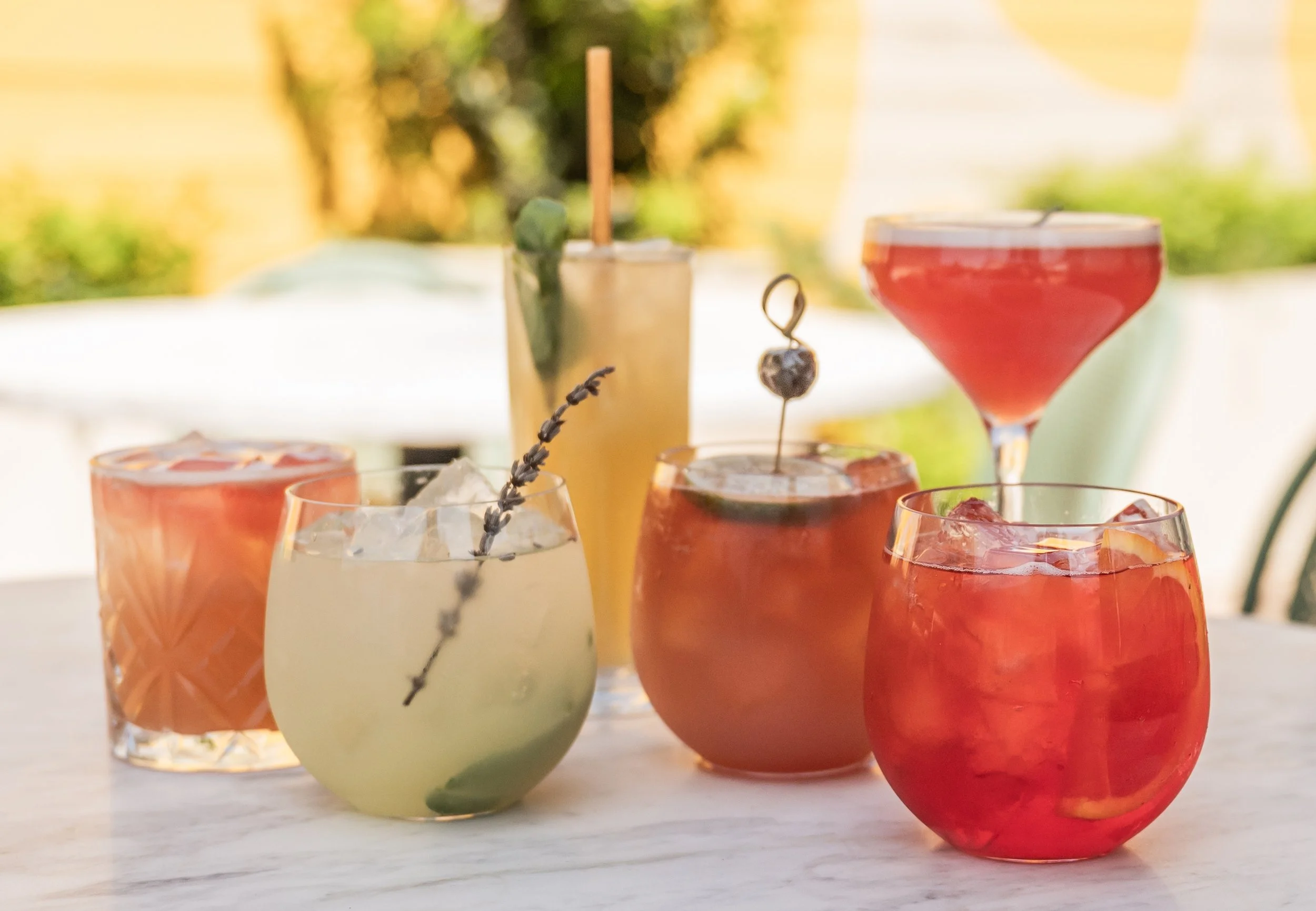 Six colorful cocktails on a white table outdoors with blurred greenery in the background.