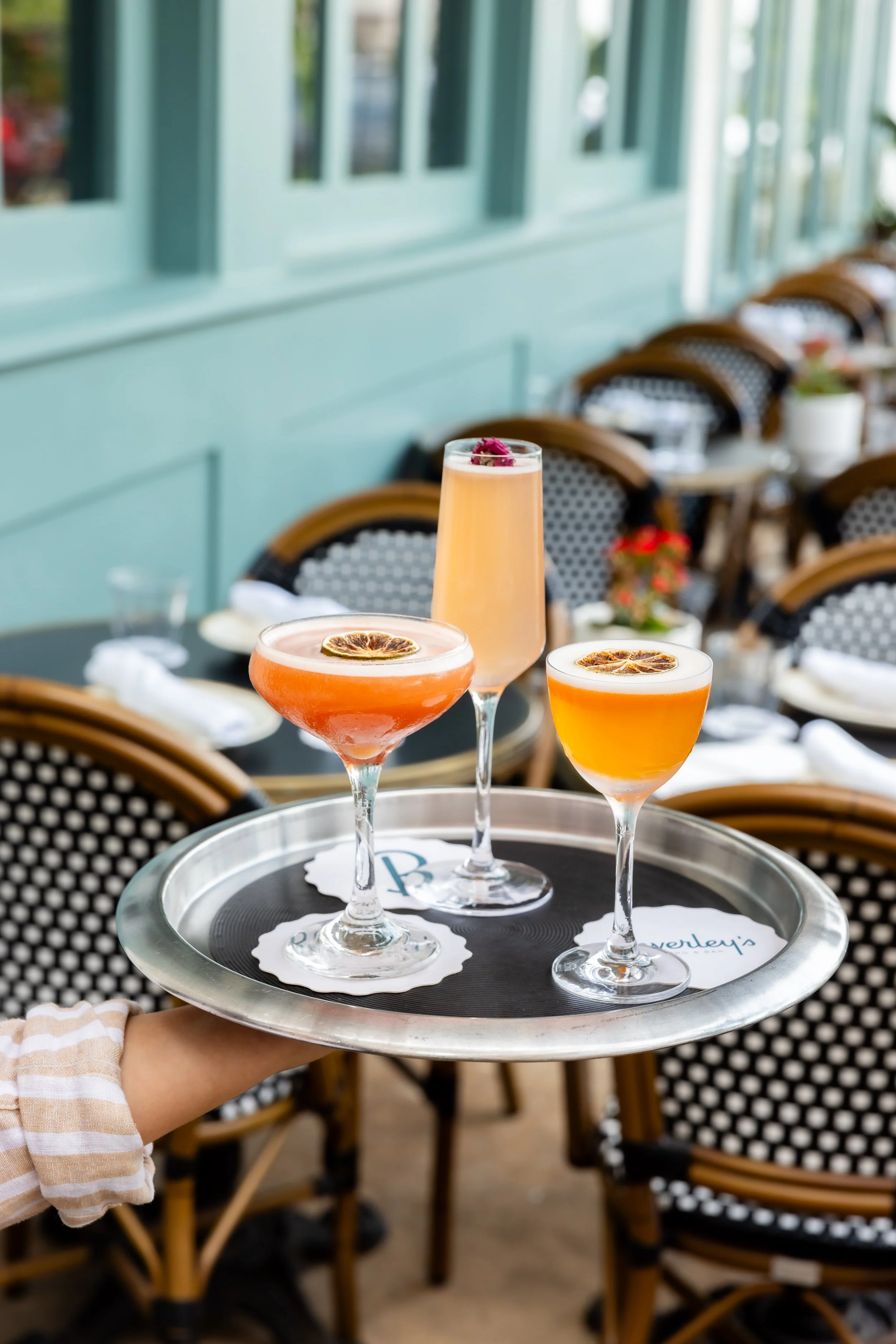 A hand holding a silver tray with three colorful cocktails garnished with dried citrus slices, set against a background of a restaurant with wicker chairs and a teal wall.