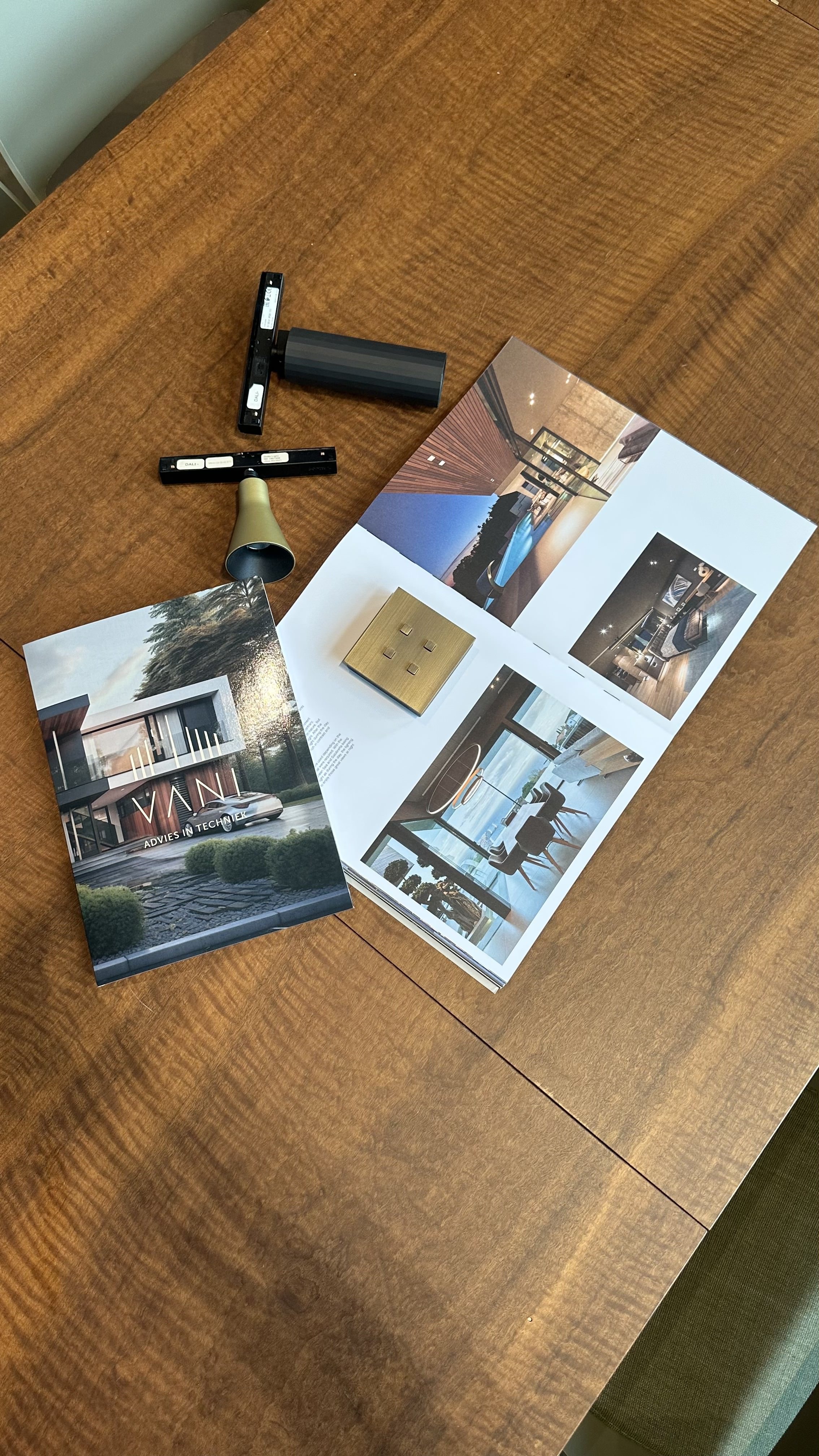 Op een houten tafel liggen brochures met interieurontwerpen, een stroomknop, een zwart component en een brandalarm.