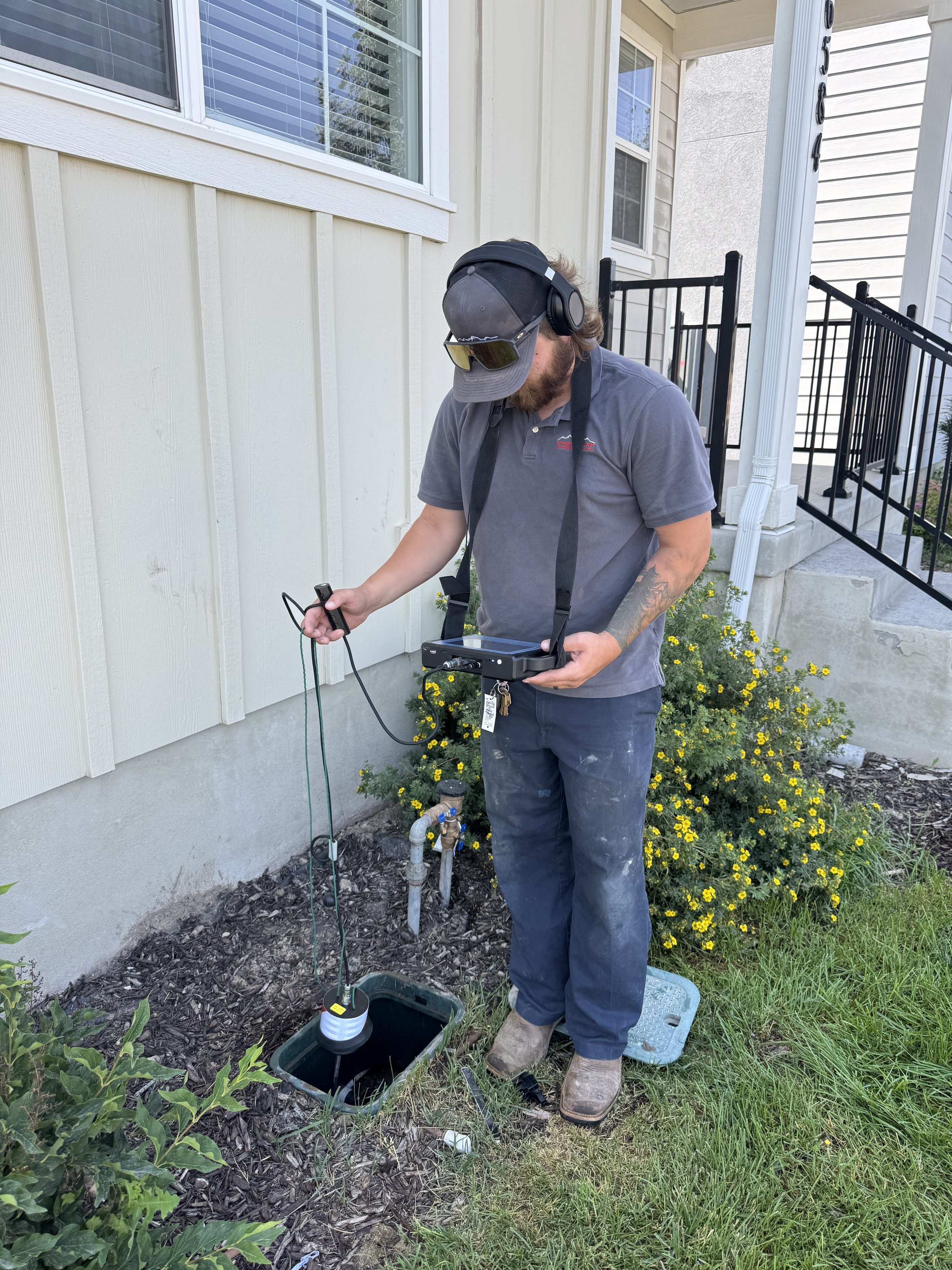 A man wearing a cap, sunglasses, headphones, and work clothes is inspecting or working on a water valve outside a house. He is holding a device connected with a cable, near an open water meter or sensor, with a storm drain nearby.