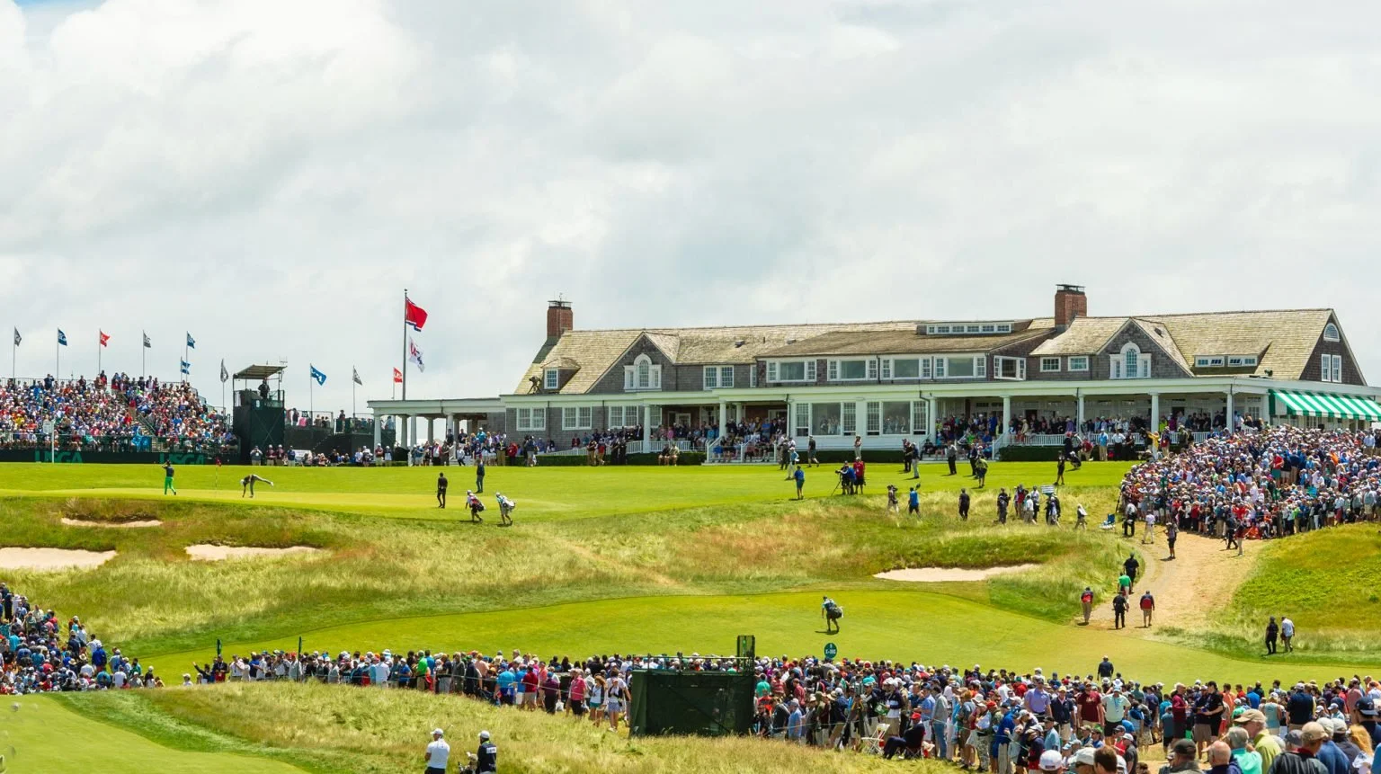 Crowd watching a golf match at a golf course, with a large clubhouse in the background.