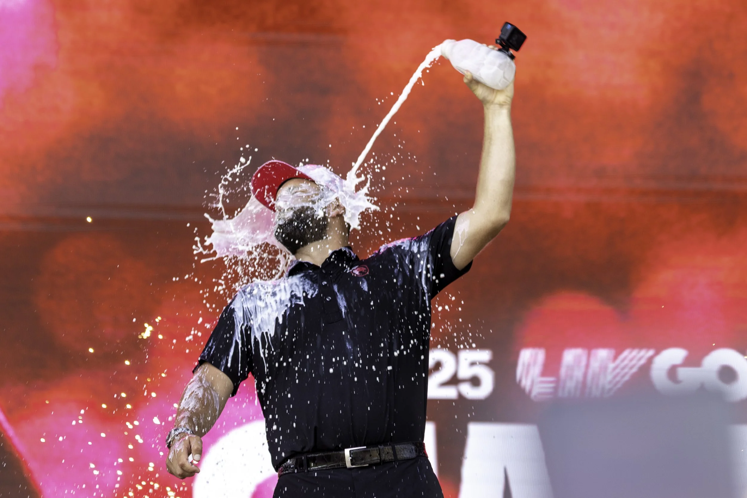 A man wearing a red cap and a black polo shirt is spraying champagne into the air at a celebration event.