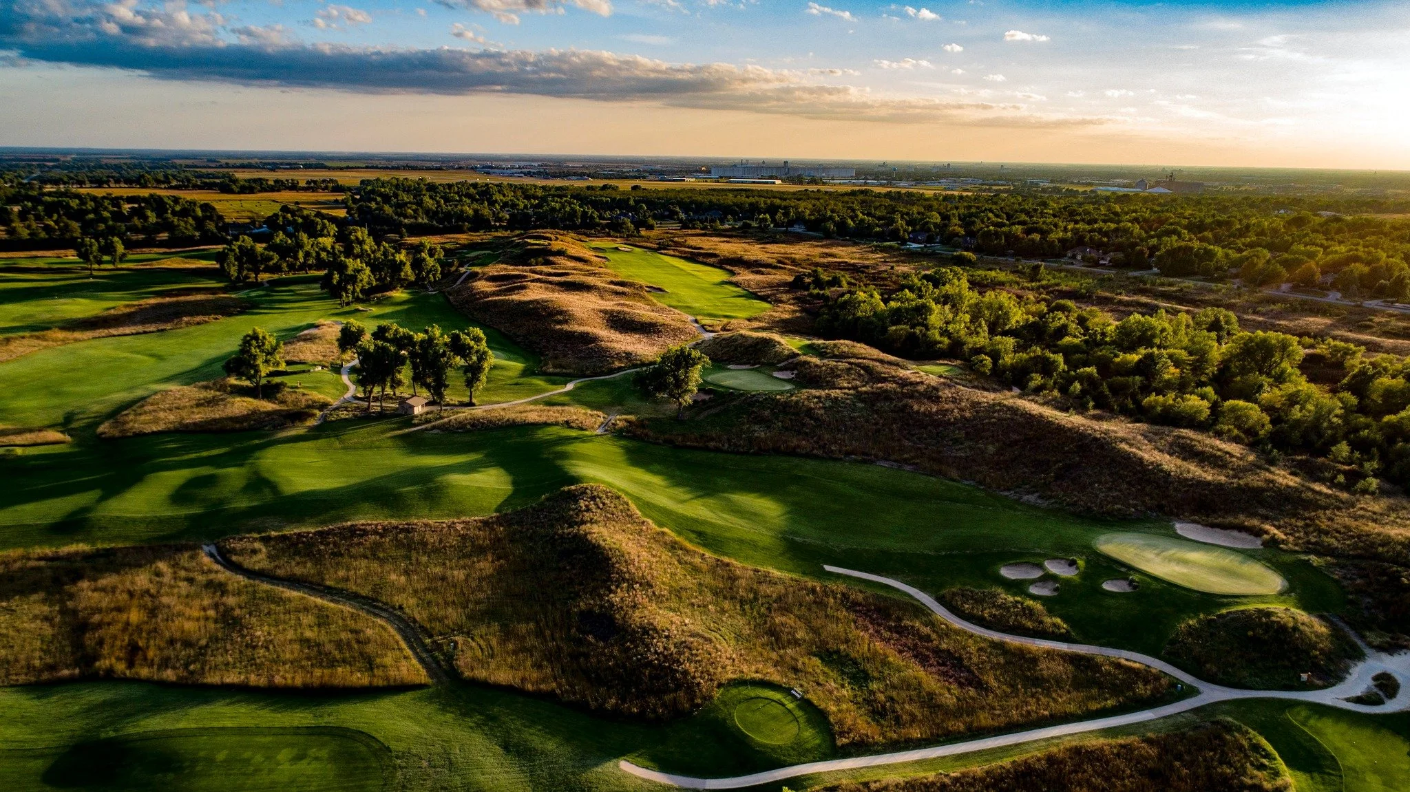 Aerial view of a golf course at sunset with green fairways, sand traps, and surrounding trees.