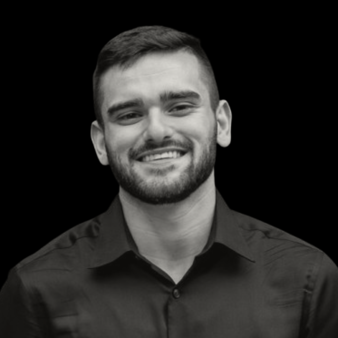 Black and white portrait of a smiling young man with short hair, a beard, wearing a collared shirt, against a plain dark background.