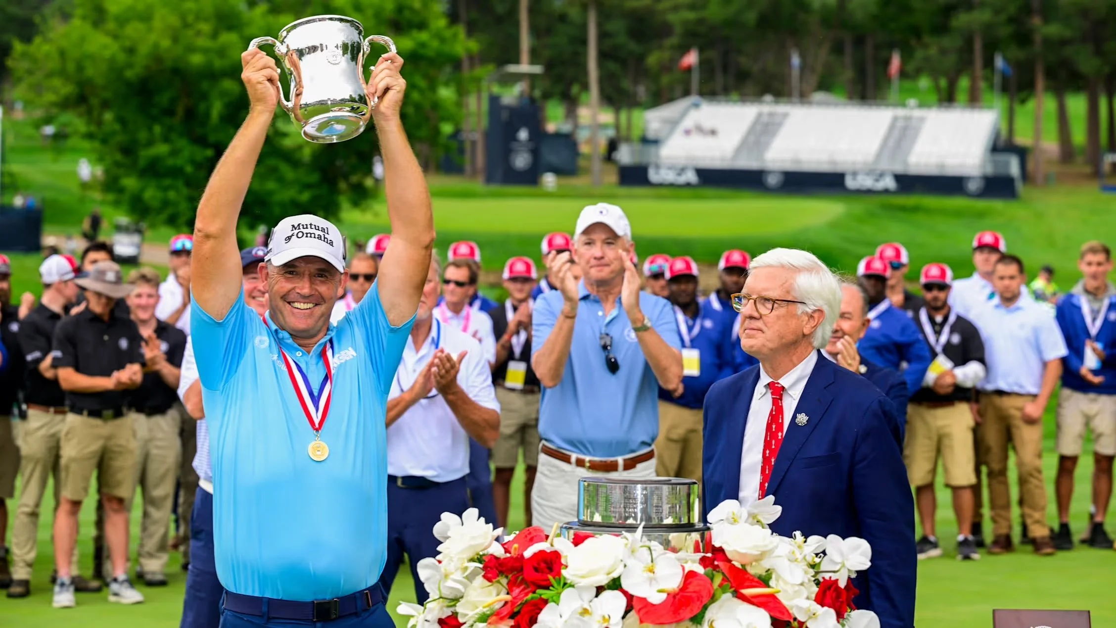 A large crowd of spectators watching a golf tournament at a golf course, with a clubhouse in the background and USGA signage on the green.