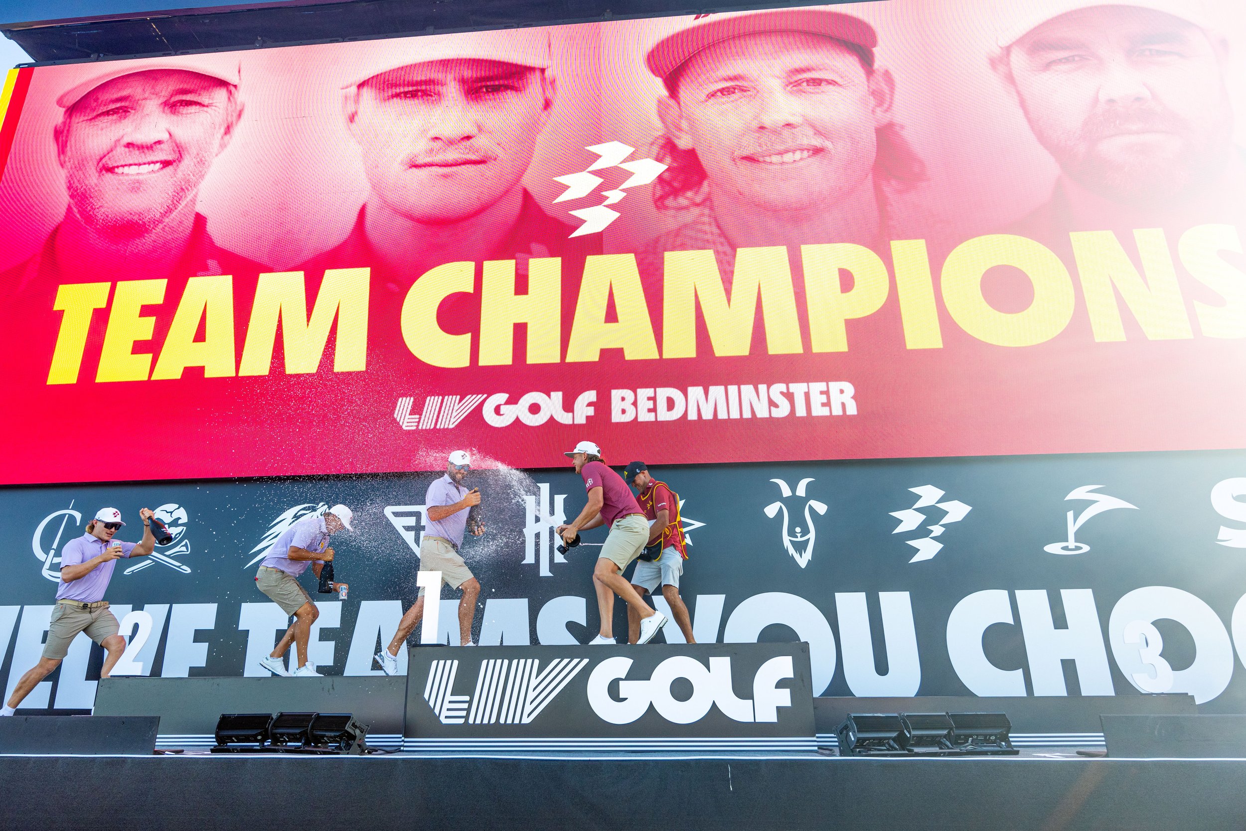 Golf players on stage celebrating team victory with large humorous backdrop showing four men's faces and the words "TEAM CHAMPIONS" with sponsor logos, some players spraying champagne.