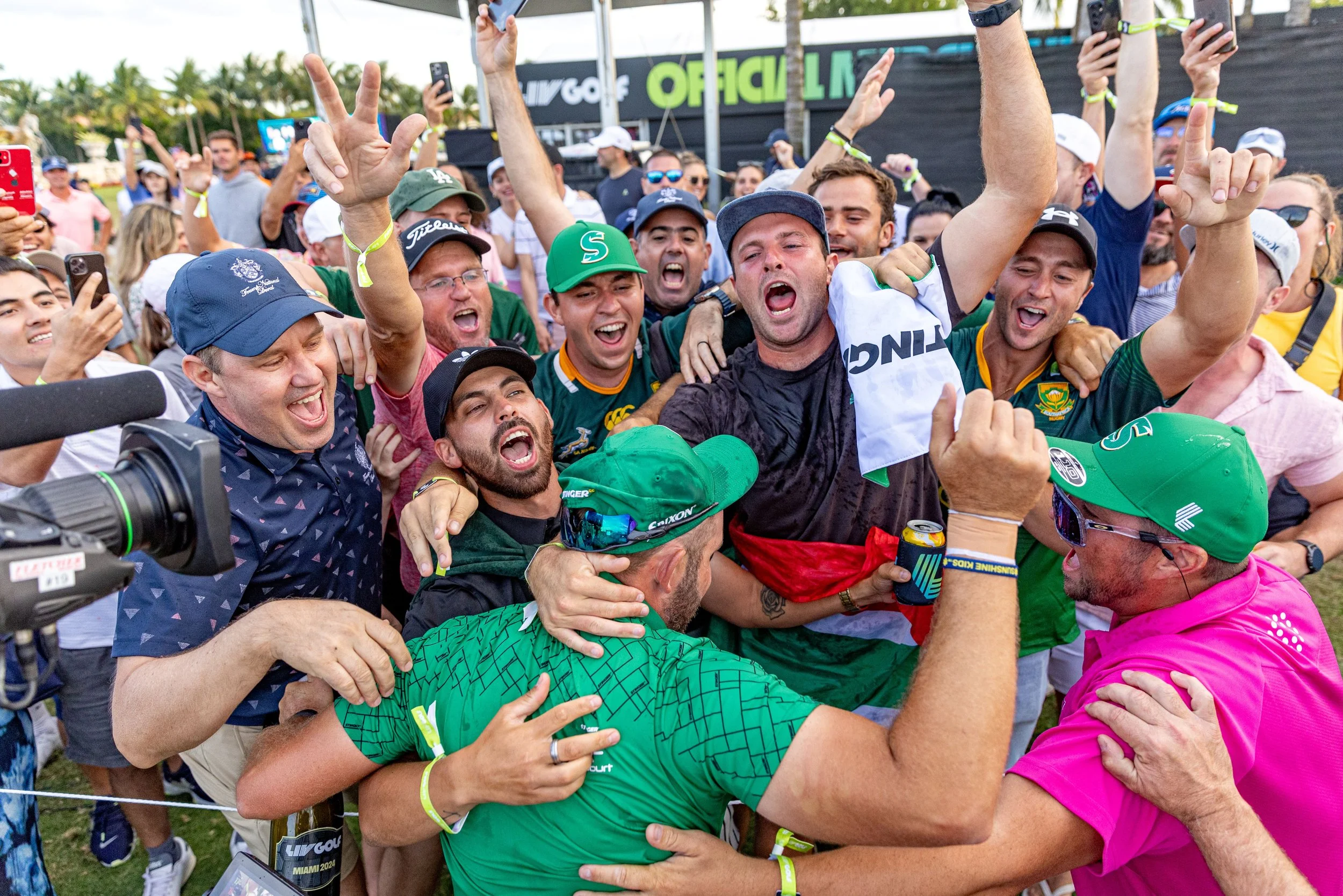 Group of people celebrating, hugging, and cheering at a sporting event, with some wearing caps and sunglasses, outdoors at a golf tournament.