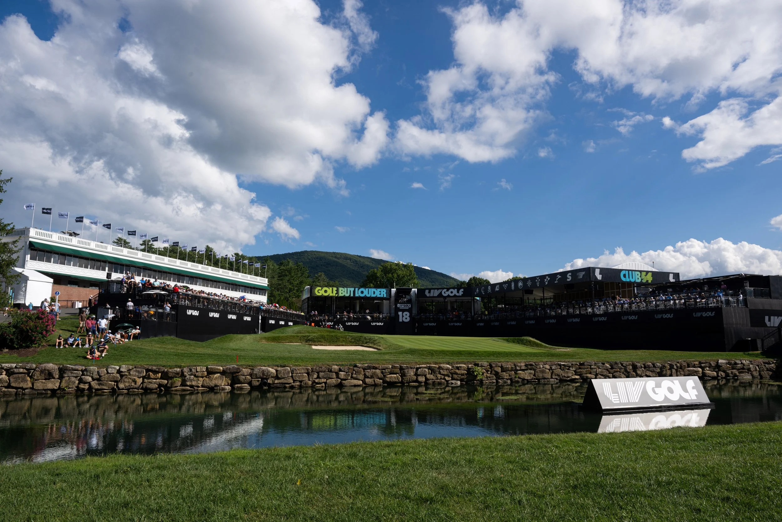 A golf tournament venue on a bright day with a large screen displaying advertising, a water hazard in the foreground, and multi-story structures with spectators and flags on top, surrounded by green hills and a partly cloudy sky.