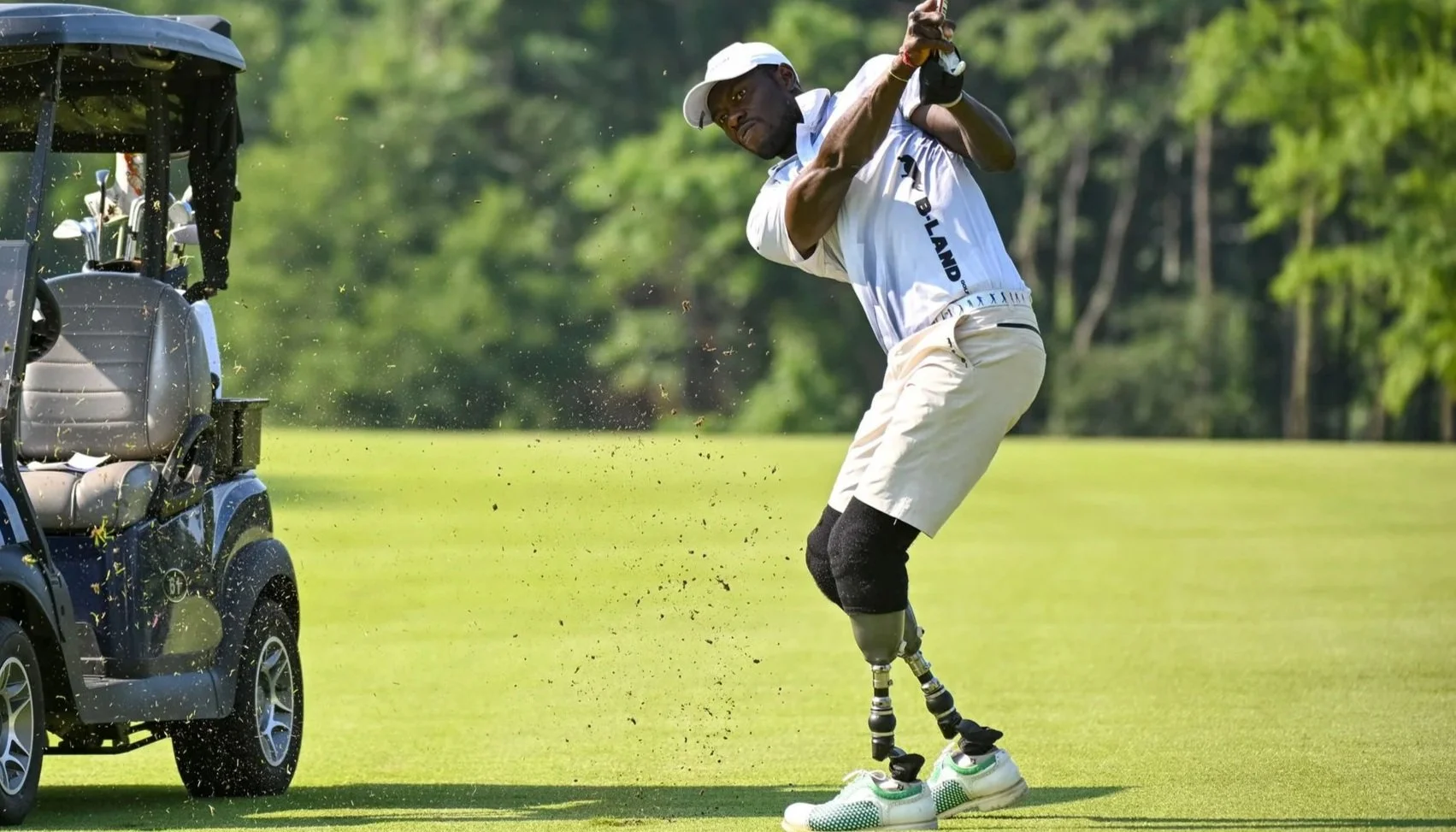 A golfer with prosthetic legs swings a golf club on the course while a golf cart is nearby, surrounded by green trees and grass.