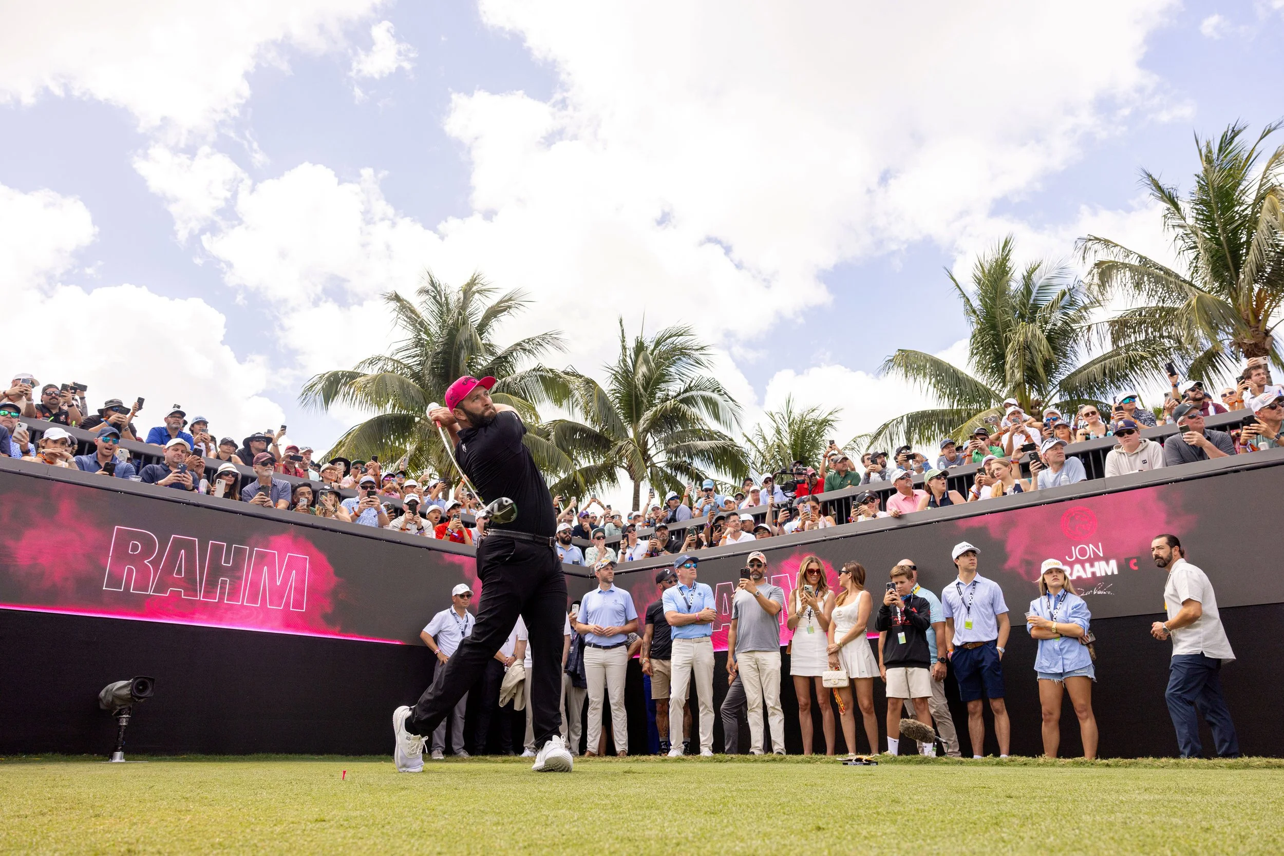 Professional golfer Jon Rahm in mid-swing during a golf tournament, surrounded by an audience on a sunny day with palm trees in the background.