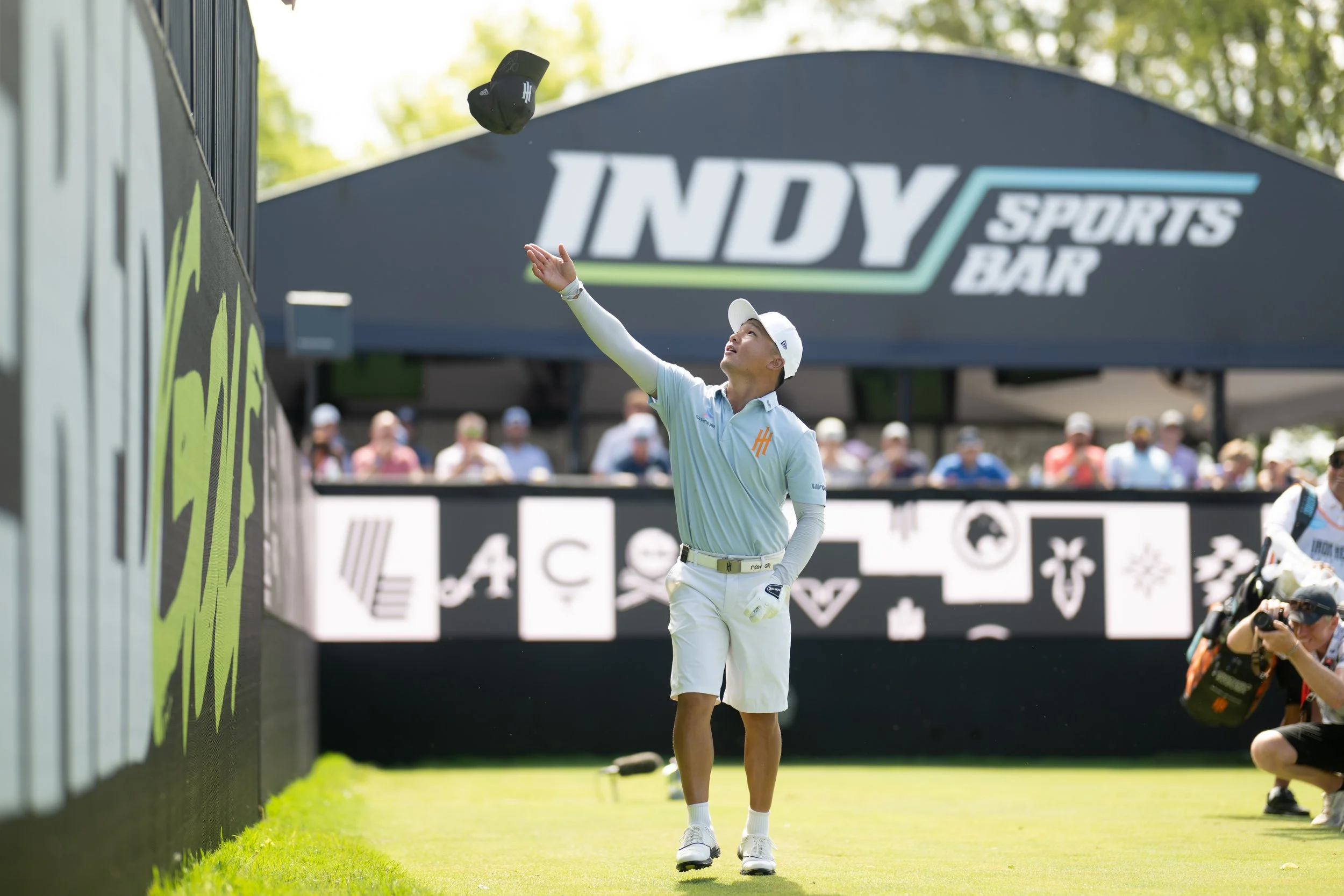 Crowd of spectators at a golf tournament, many wearing sunglasses and hats, on a sunny day near the golf course.