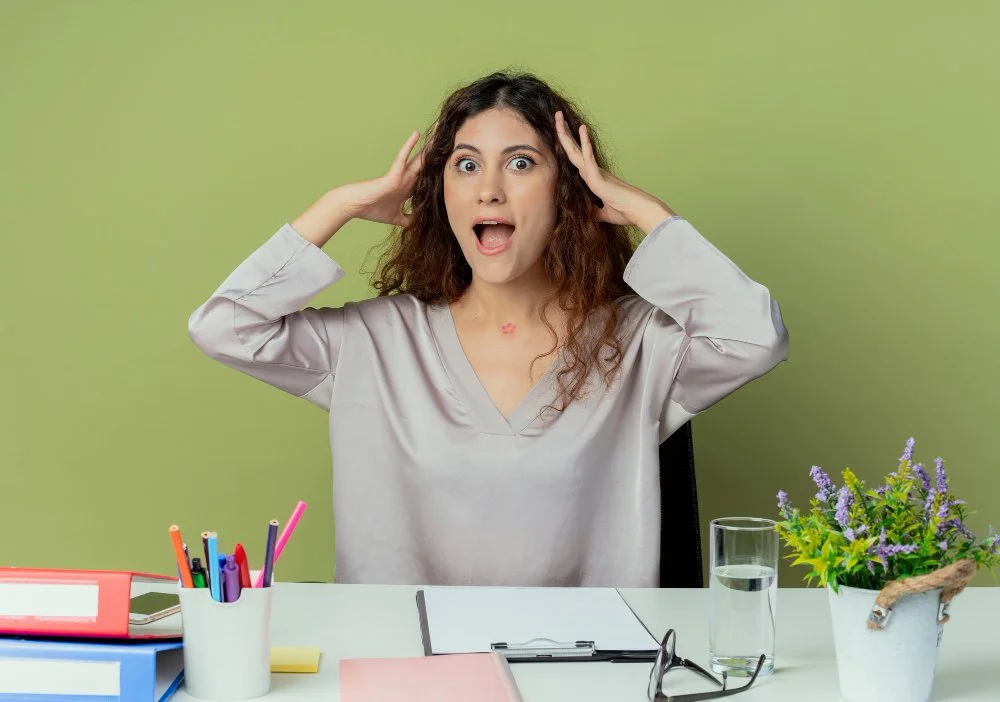 A woman sitting at a desk with a surprised or overwhelmed expression, holding her head with her hands, with office supplies and a potted plant on the desk.