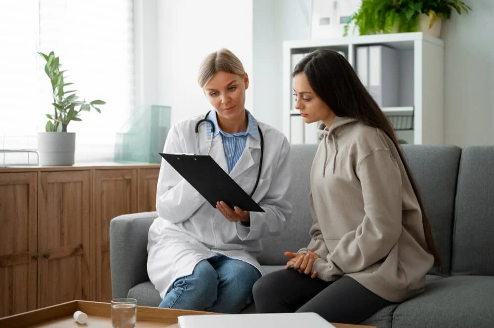 A female doctor explaining something on a clipboard to a young woman sitting on a sofa in a bright, modern clinic or hospital room.